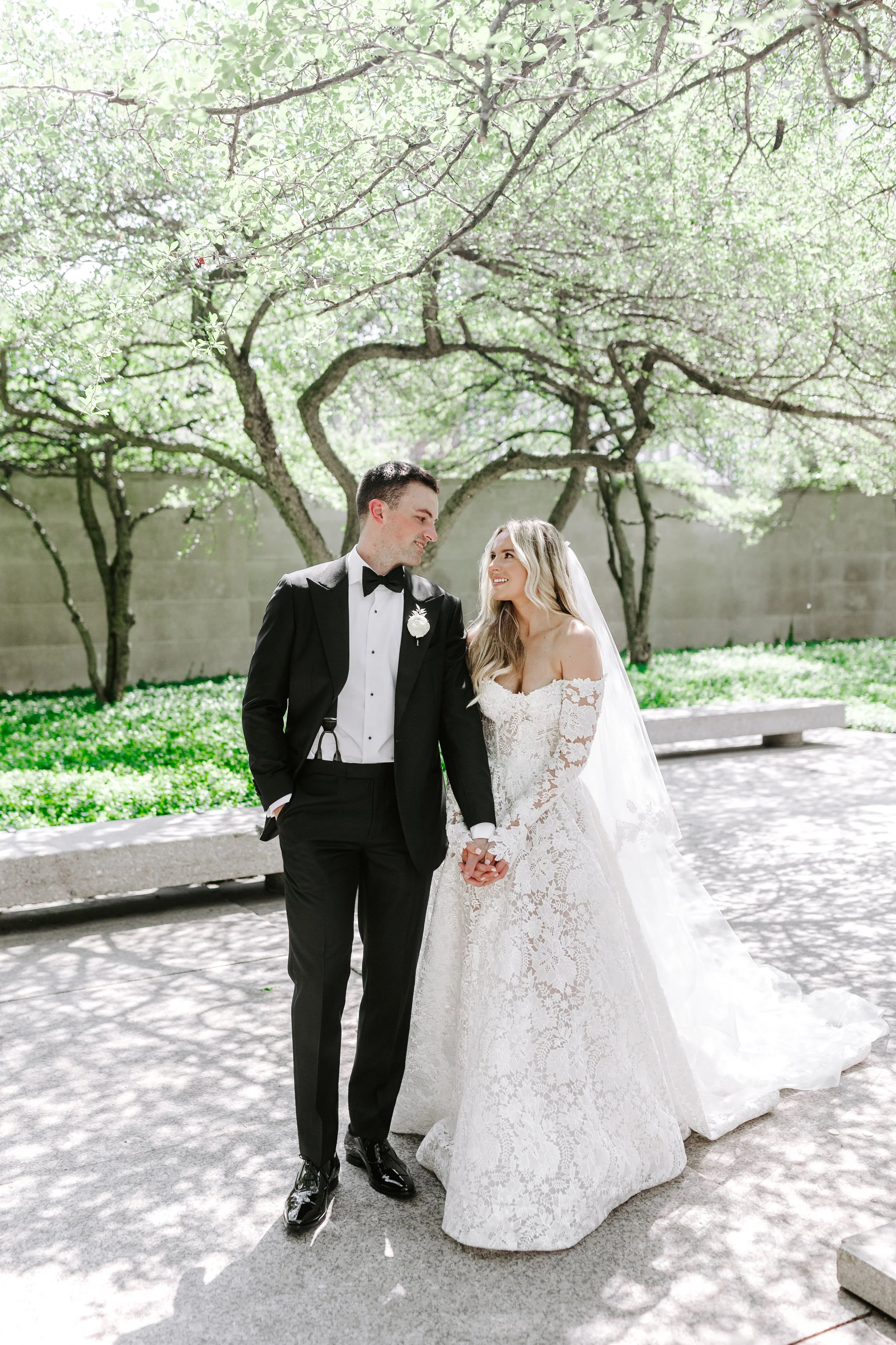 A bride and groom holding hands and gazing at each other outdoors under a leafy tree on a sunny day.