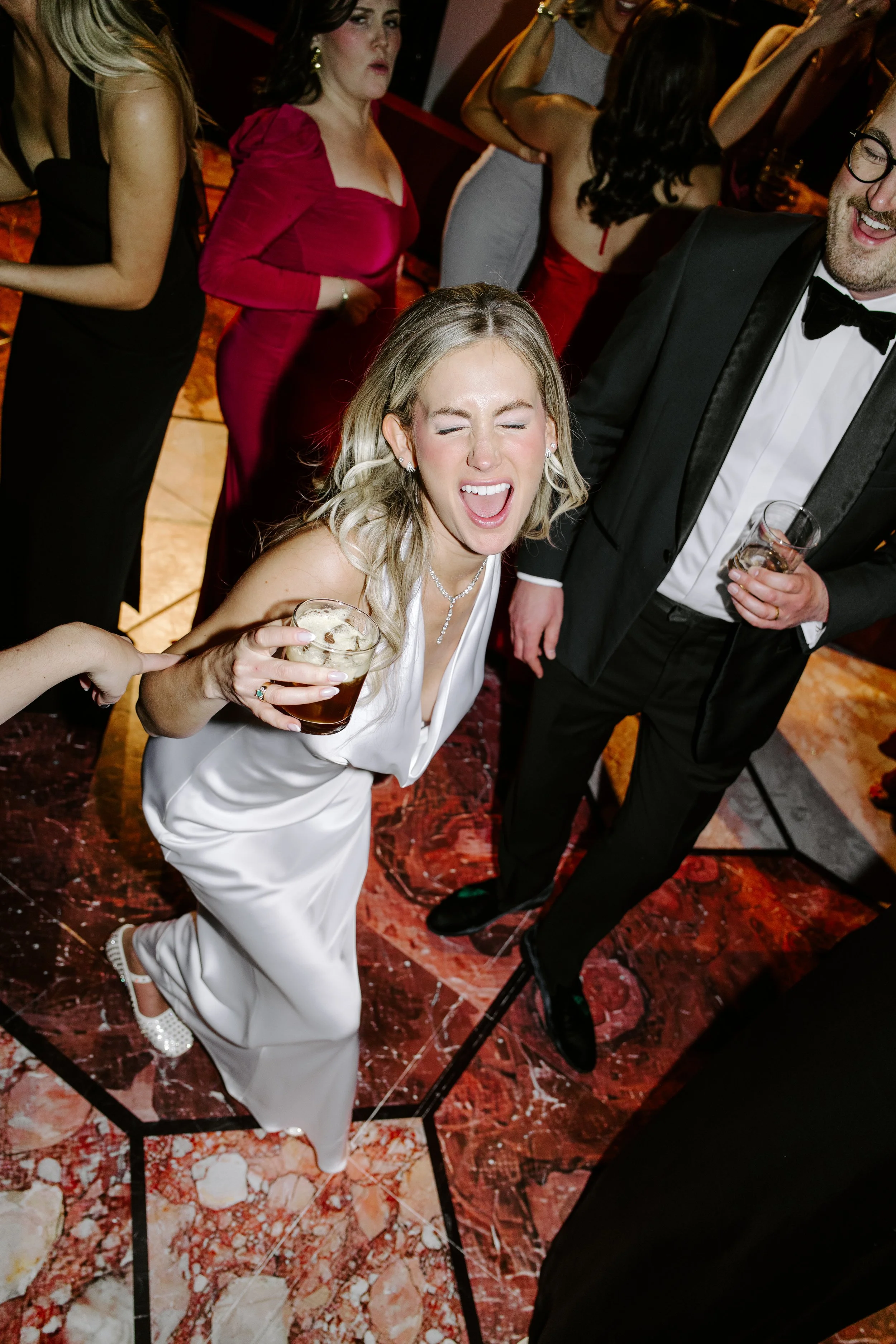 Group of people dancing and socializing at a party or celebration. Woman in white dress holding a drink, laughing with eyes closed, surrounded by others in formal attire.