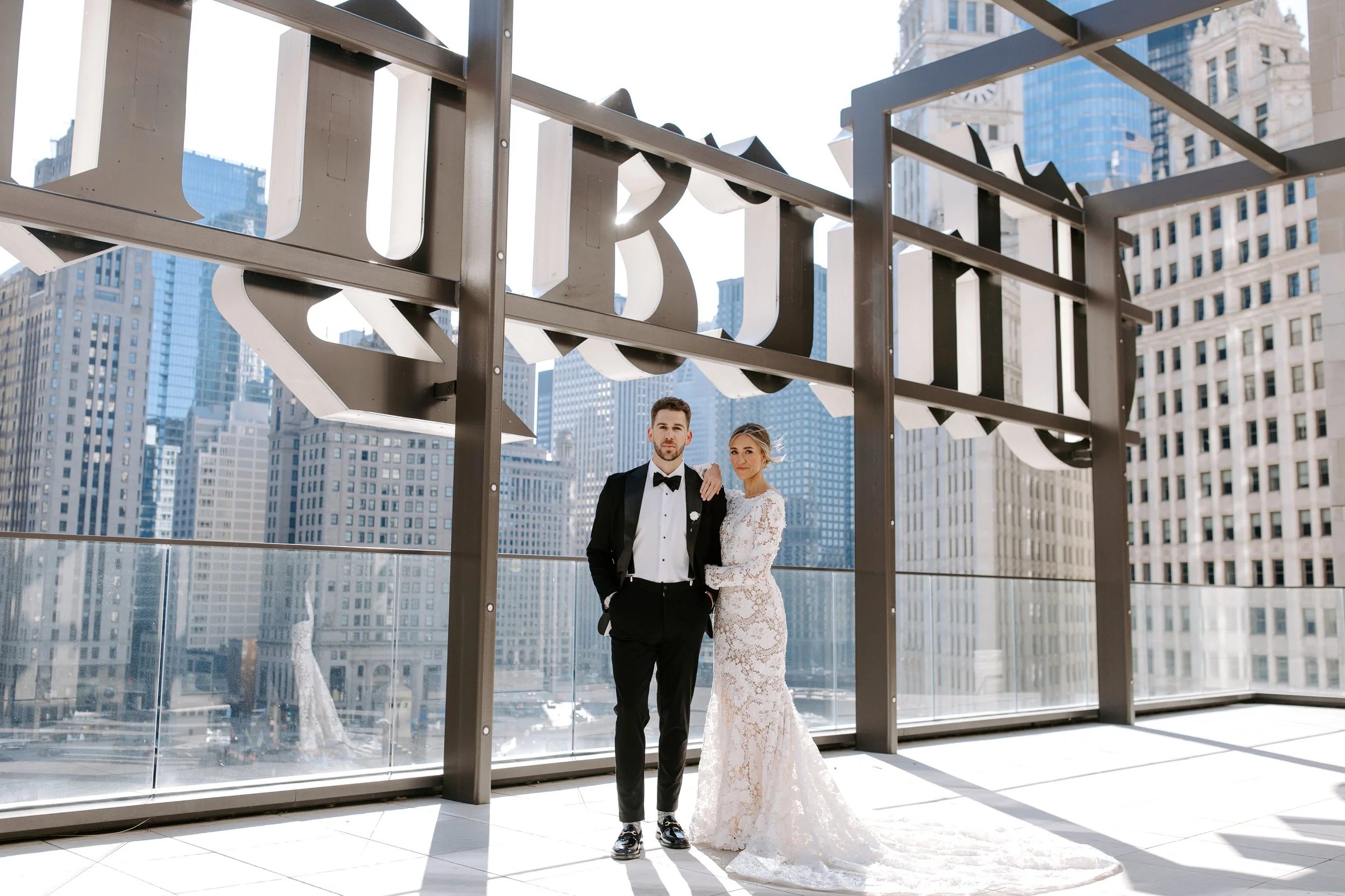 A bride and groom in formal wedding attire standing close together on a rooftop with tall city buildings in the background.