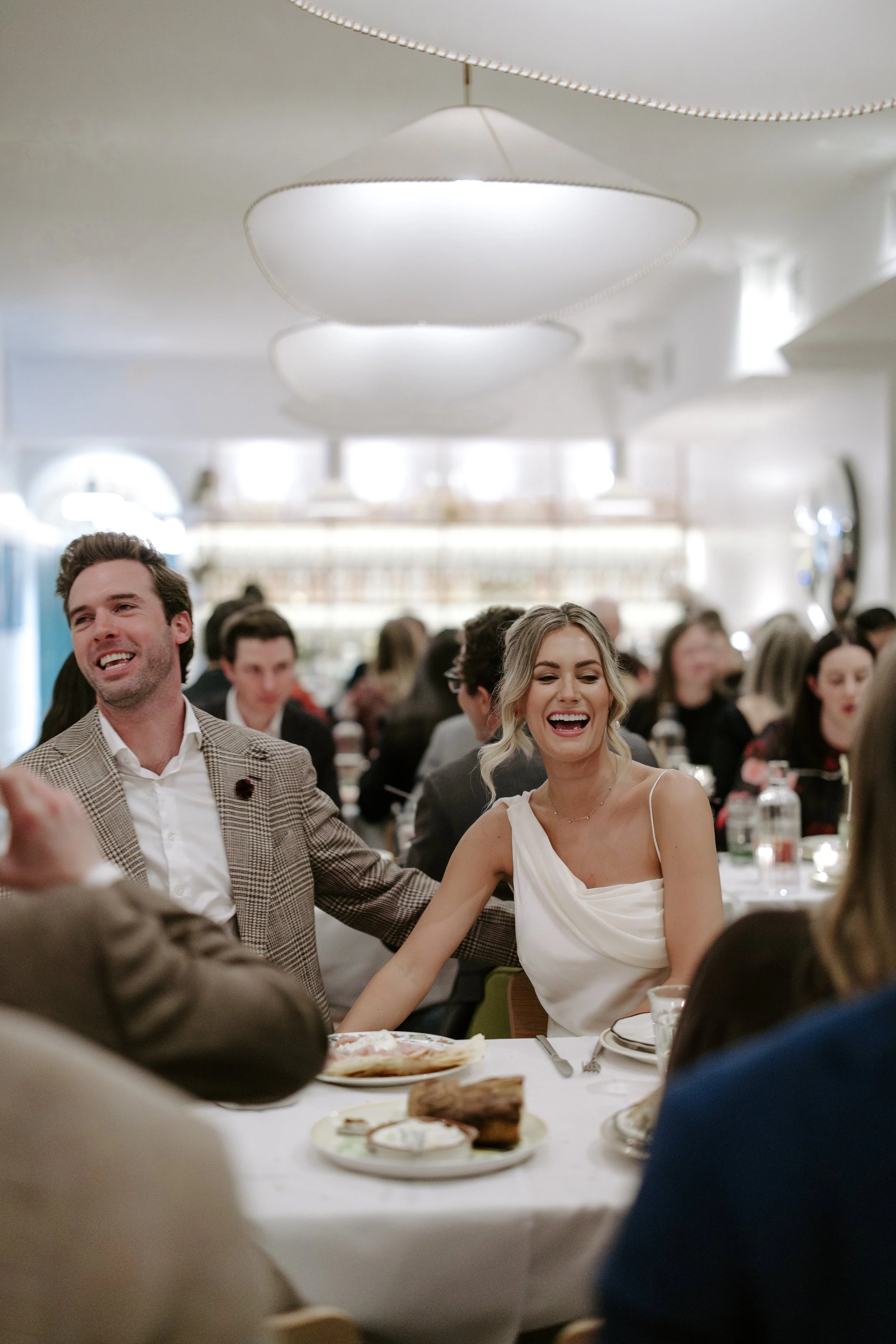 A group of people enjoying a meal at a formal event, with a man and woman smiling and holding hands, surrounded by other guests, in a well-lit banquet hall with modern lighting fixtures.