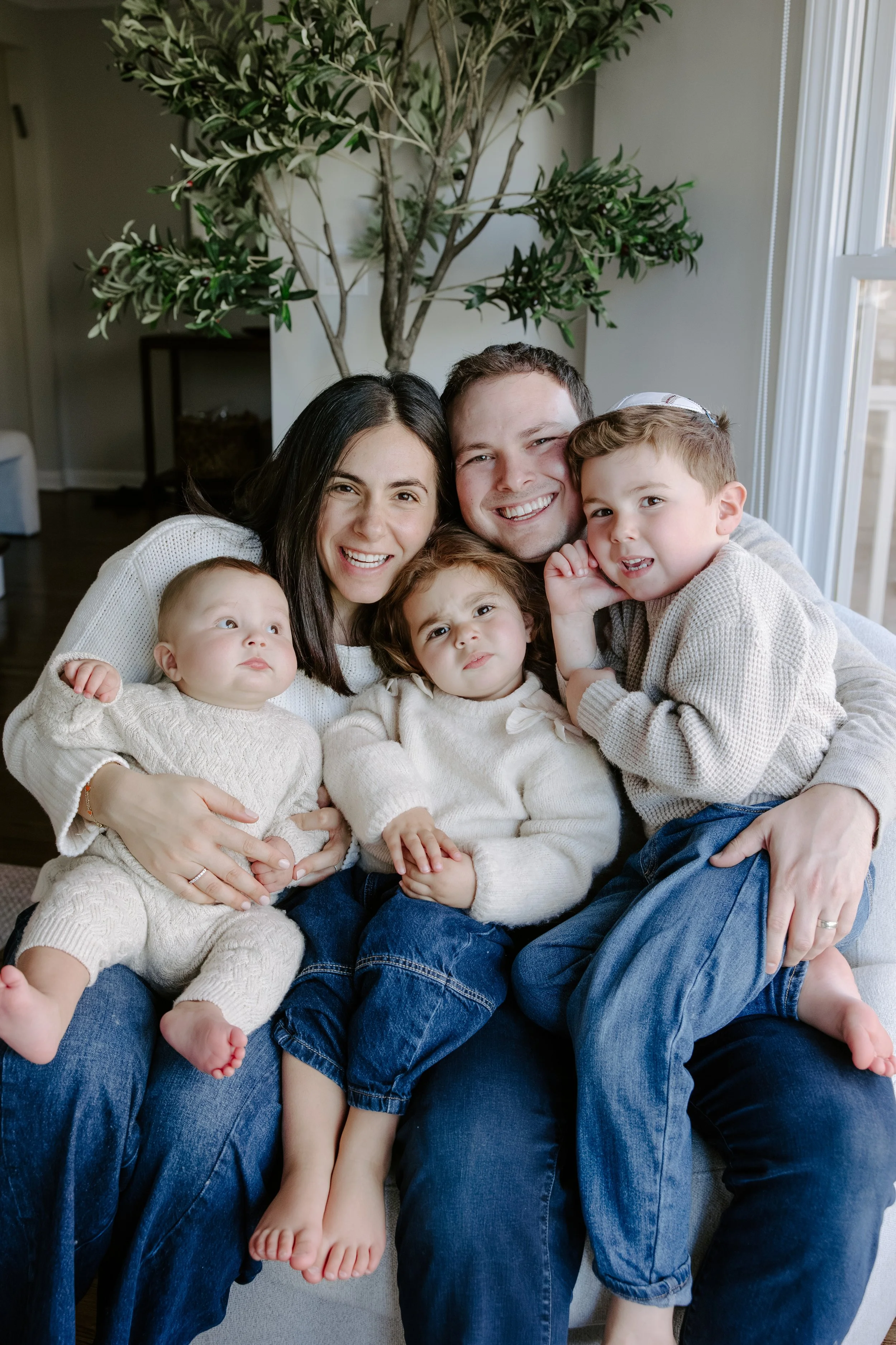 A happy family of five, including two adults and three children, sitting together on a couch in a living room with natural light and a large indoor plant in the background. The family is smiling and cuddling.