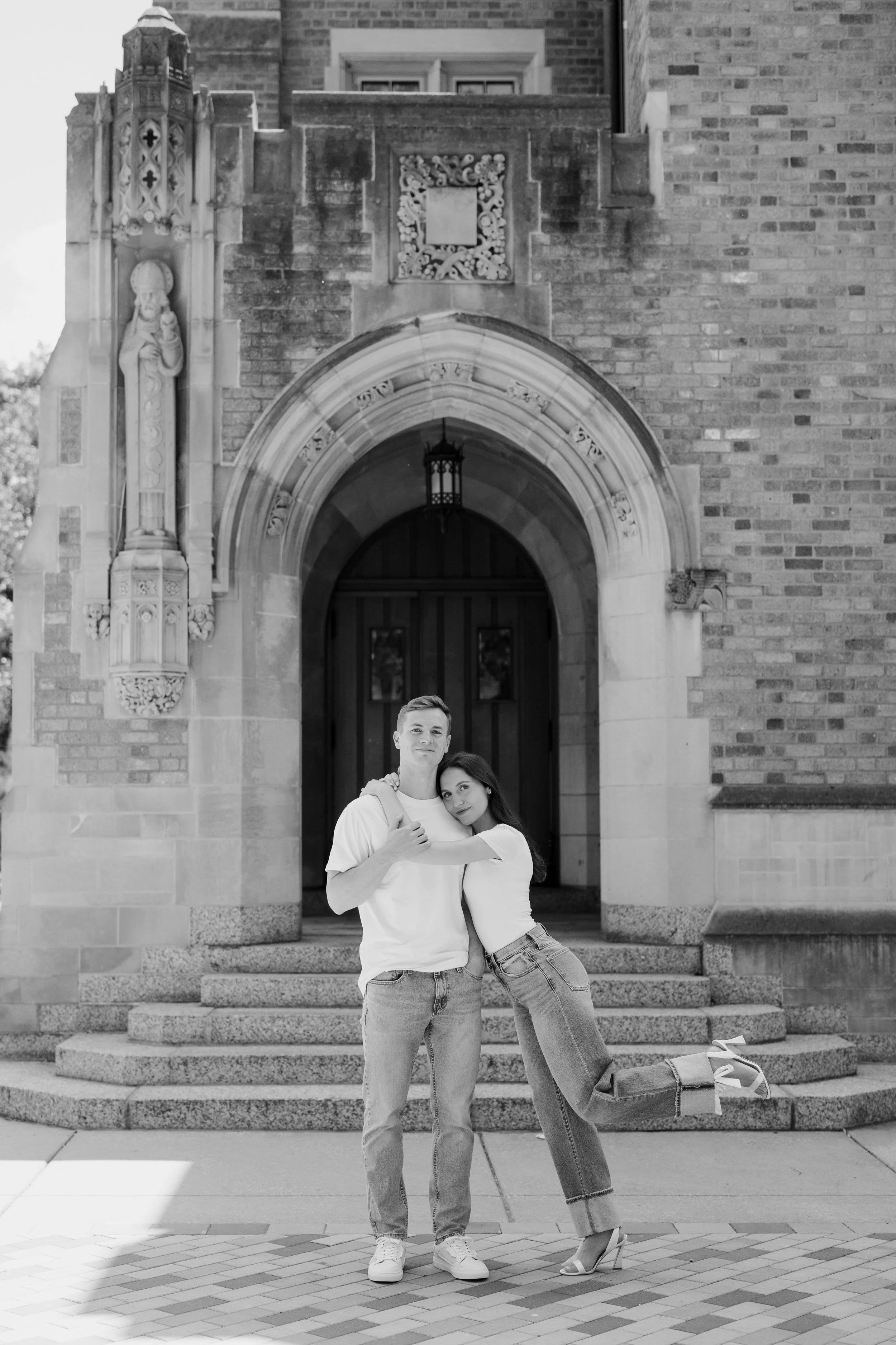 A young man and woman embrace and pose playfully on the steps of a historic building with detailed stonework, including a statue of a religious figure on the left side and an ornate archway above the entrance.