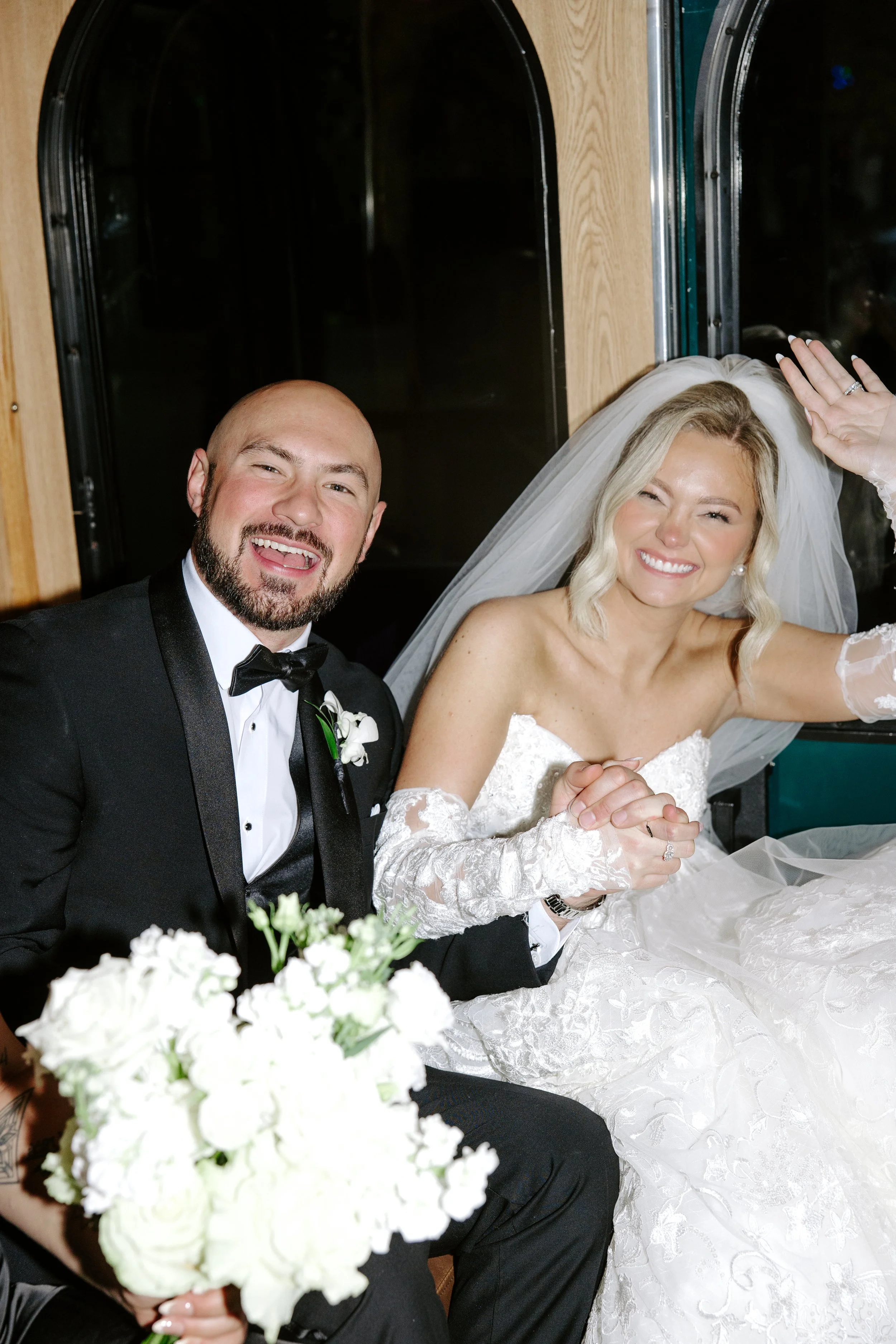 A happy bride and groom at their wedding, sitting together with the bride smiling and the groom smiling with his tongue out, holding hands, with white flowers in the foreground.