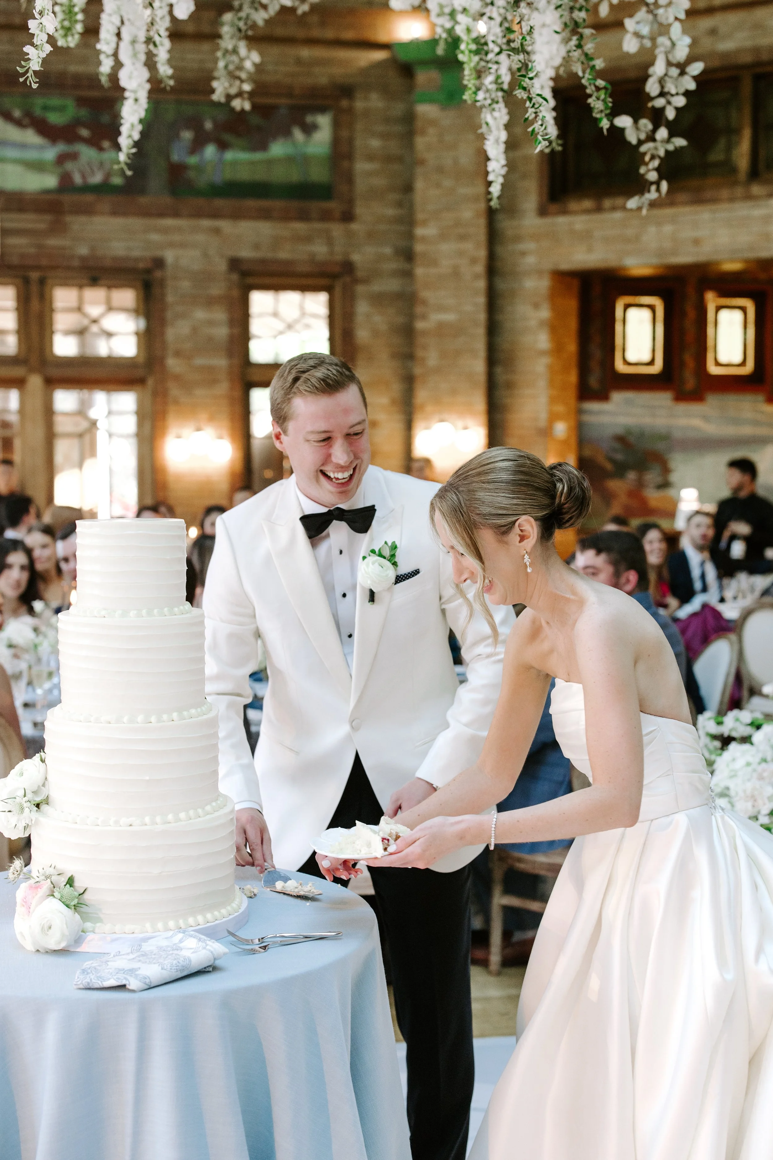 A newlywed couple cutting a wedding cake at their reception. The bride in a strapless wedding dress, the groom in a white tuxedo with a black bow tie. The cake is white, multi-tiered with decorations of white flowers. The setting is a rustic hall wit