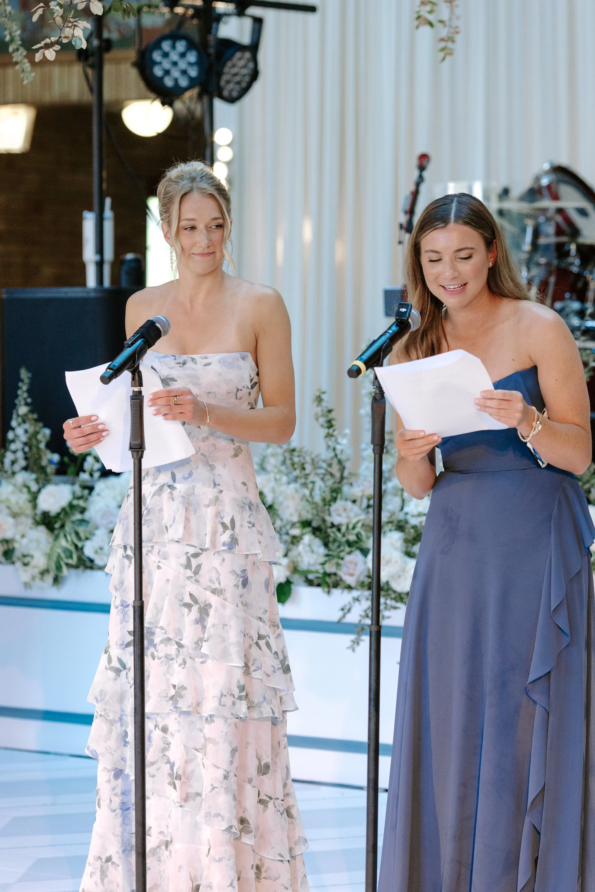 Two women in formal dresses standing behind microphones at an indoor event, with one reading from a paper and the other looking at her, surrounded by floral decorations and stage lighting.