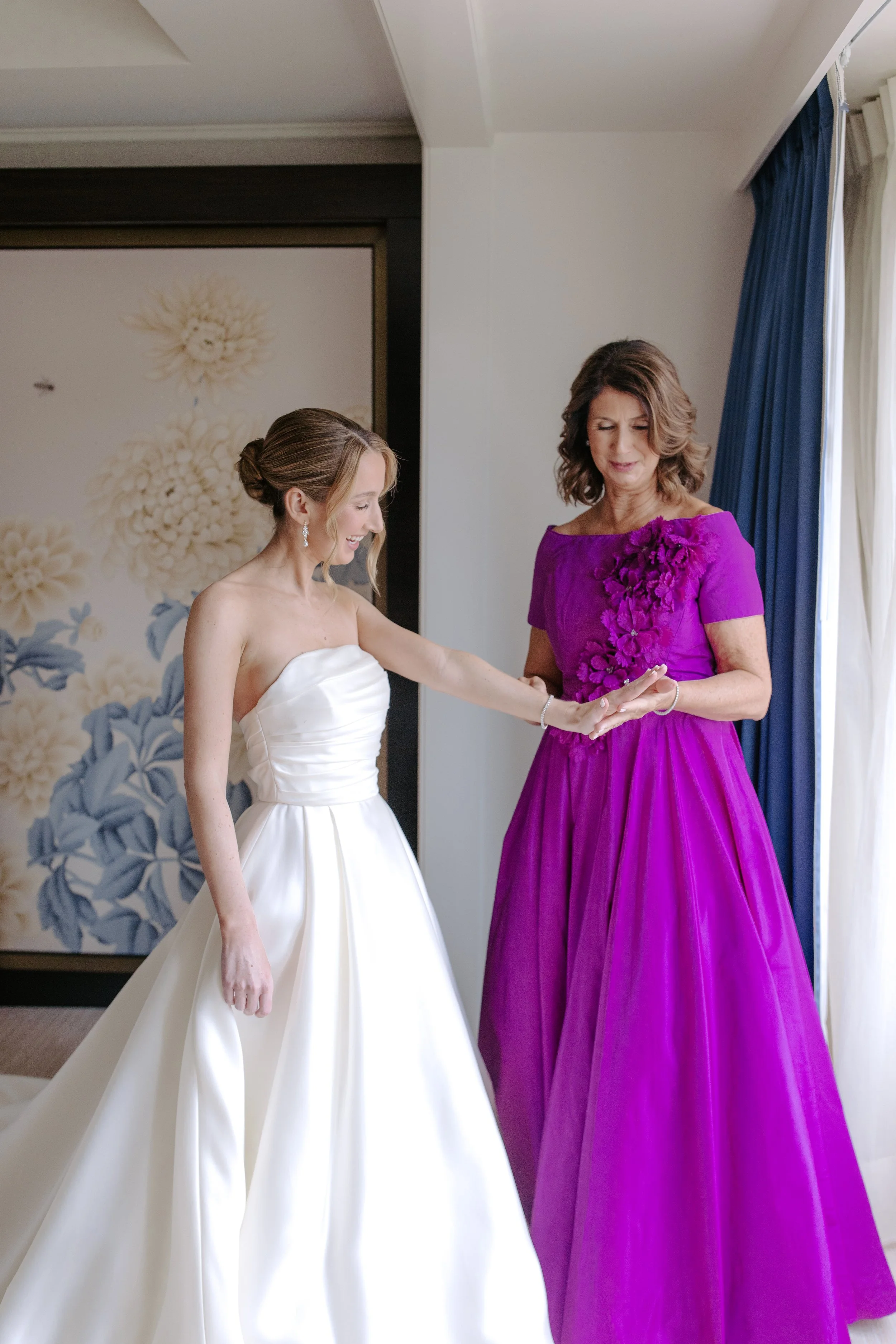 A bride in a white wedding gown holding hands with a woman in a purple dress, inside a room with floral wallpaper and blue curtains.