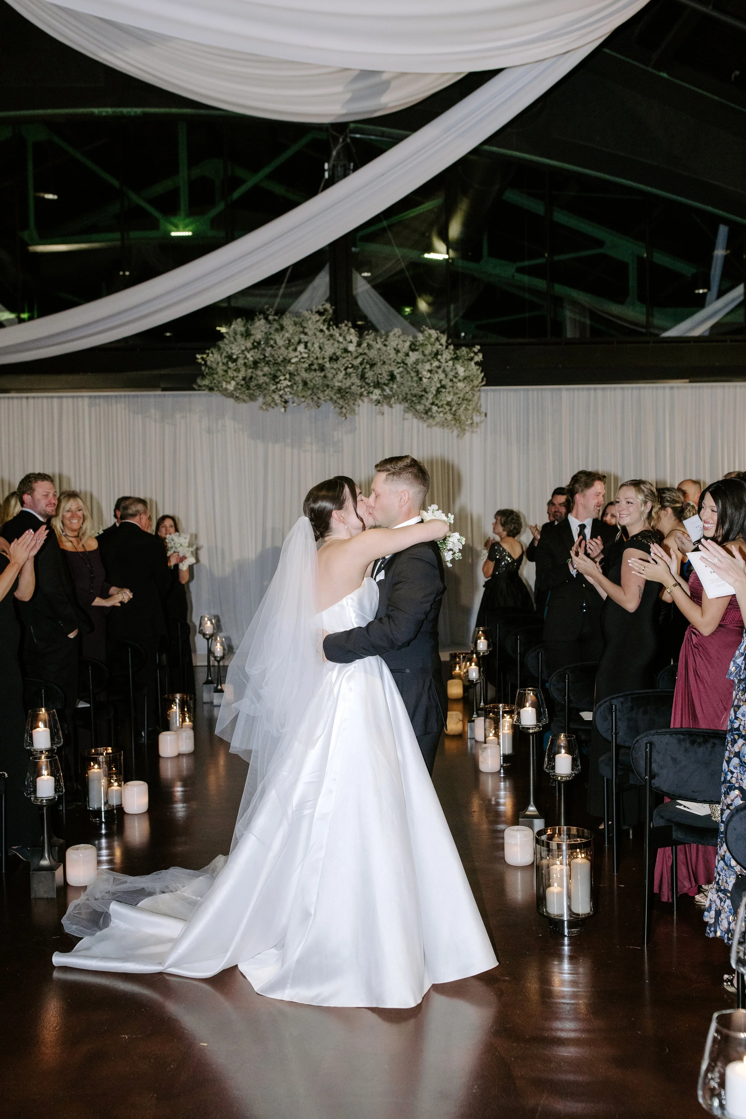 Couple sharing their first dance at a wedding reception, surrounded by guests clapping and smiling, with candles on the floor and a decorative floral arrangement above them.