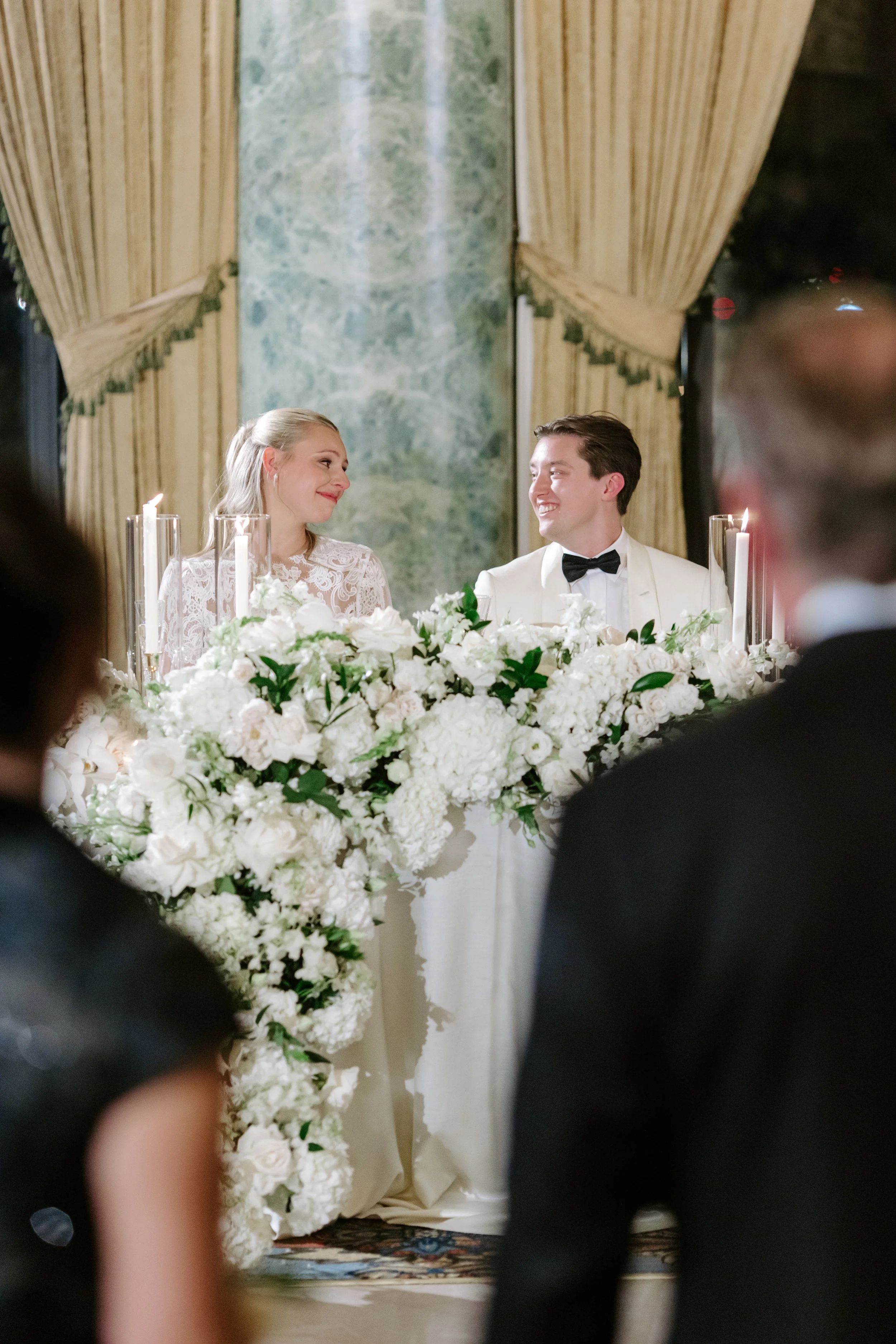 A bride and groom sitting at a wedding reception table decorated with white flowers and candles, smiling at each other, with elegant curtains and a green marble wall in the background.