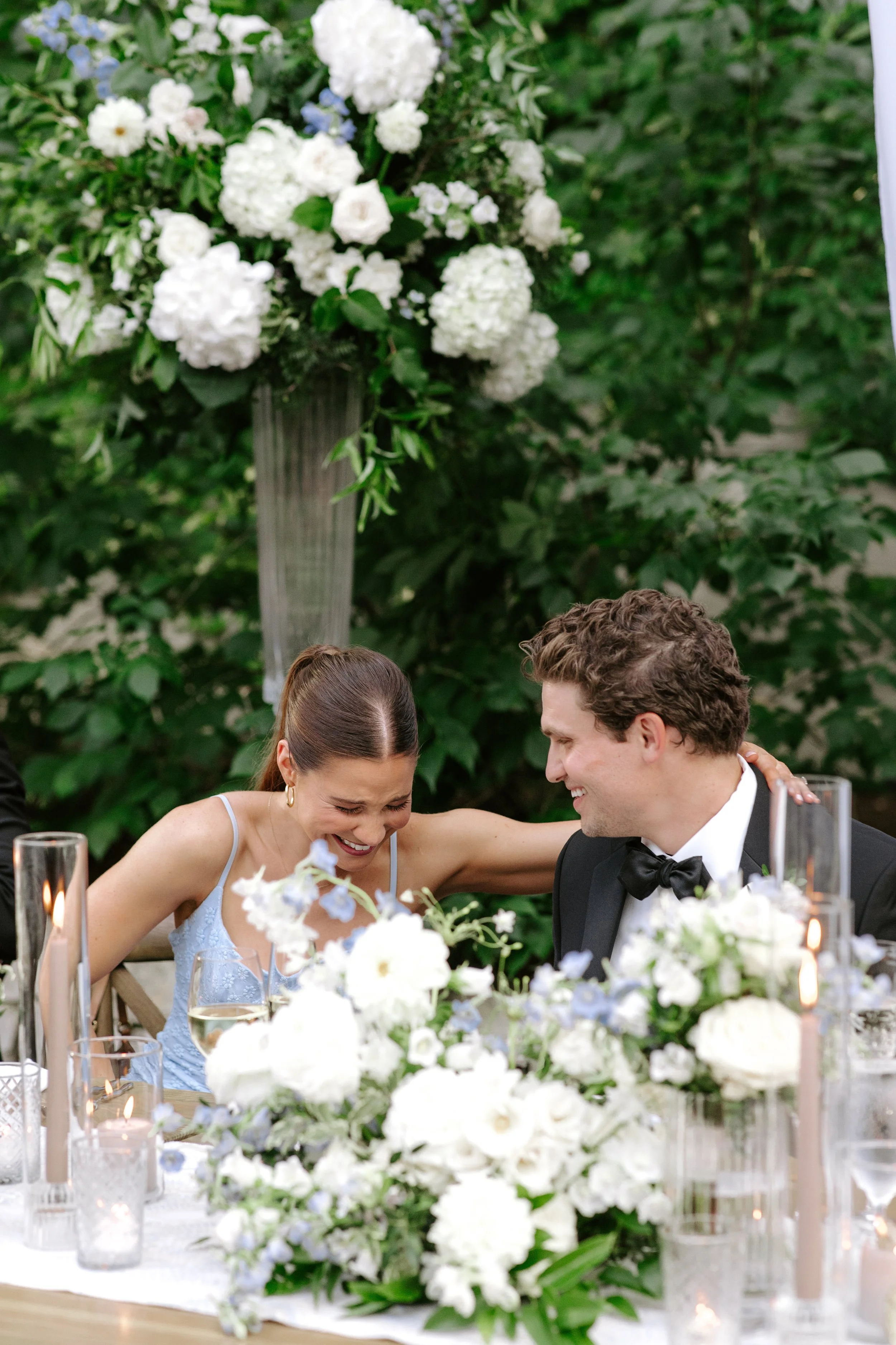 A bride and groom sitting at a wedding reception table, smiling and leaning towards each other, surrounded by floral arrangements and candles.