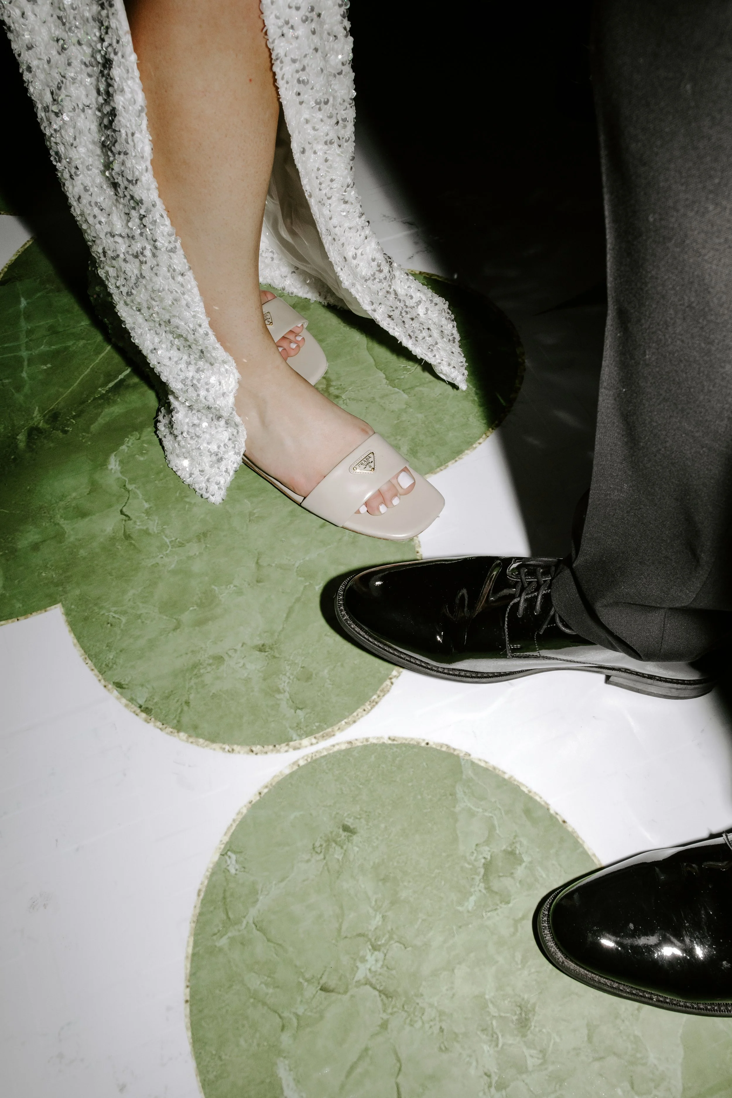 Close-up of two pairs of legs and feet, one in a white beaded dress with open-toe heels, the other in black dress shoes, standing on a green and white circular patterned floor.