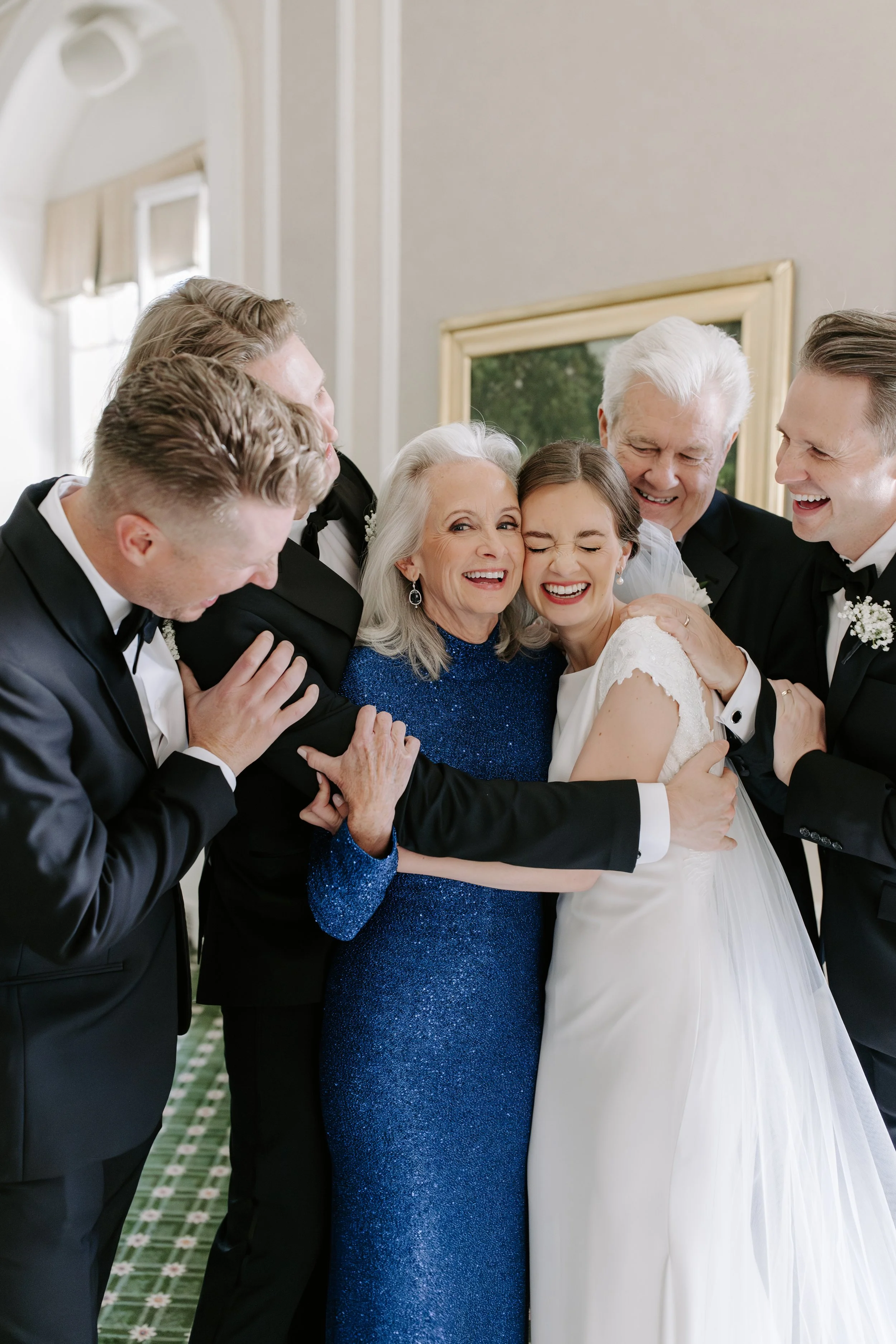 A group of people hugging and laughing at a wedding, including a bride in a white gown and an older woman in a blue dress.