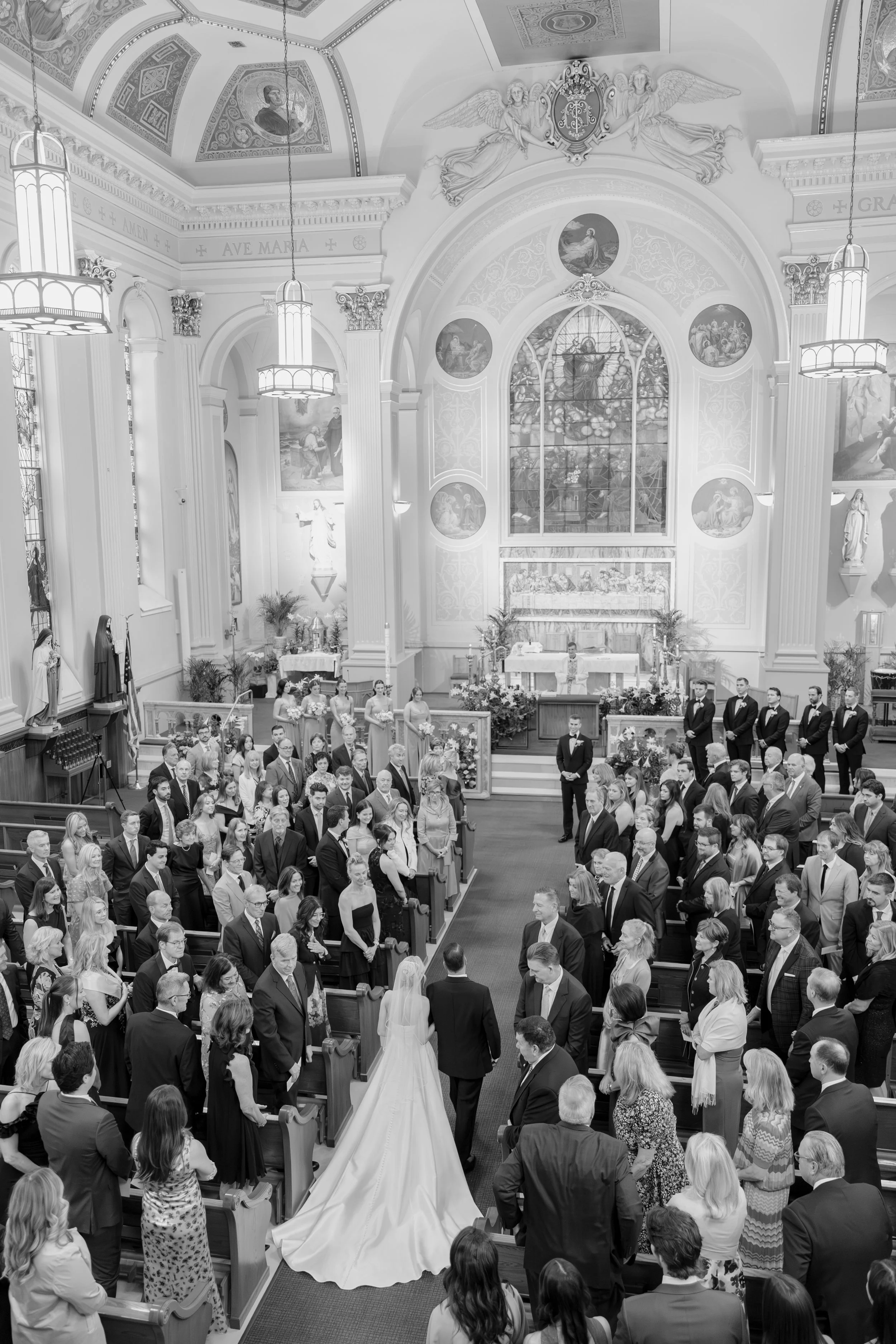 A wedding ceremony inside a church with many guests, a bride in a white gown, and a groom in a suit, with ornate stained glass windows and religious artwork in the background.