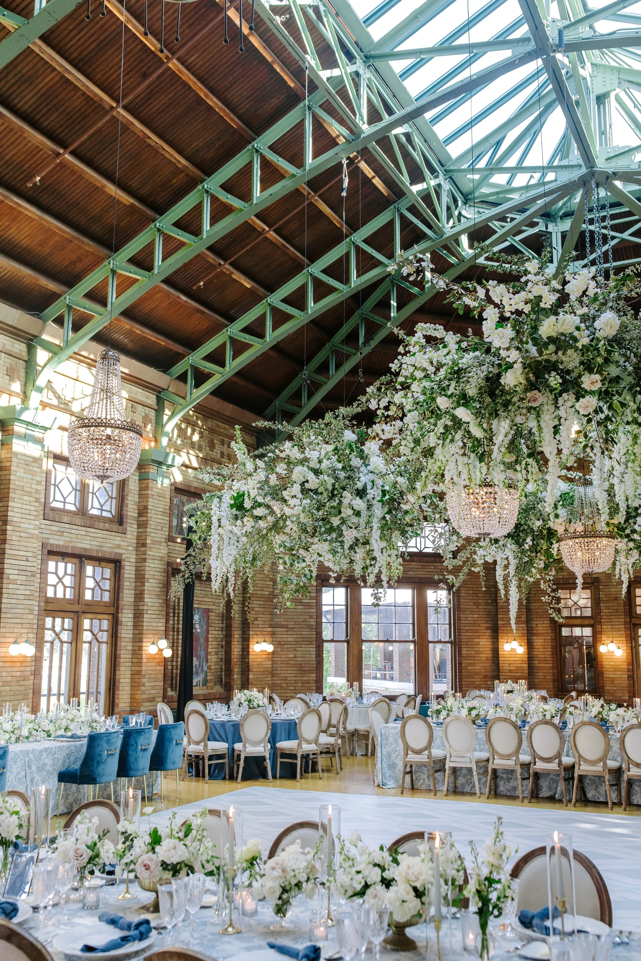 Elegant event hall with large floral arrangements, round tables with white tablecloths, and a green industrial-style ceiling with wooden accents and hanging chandeliers. There are large windows letting in natural light.