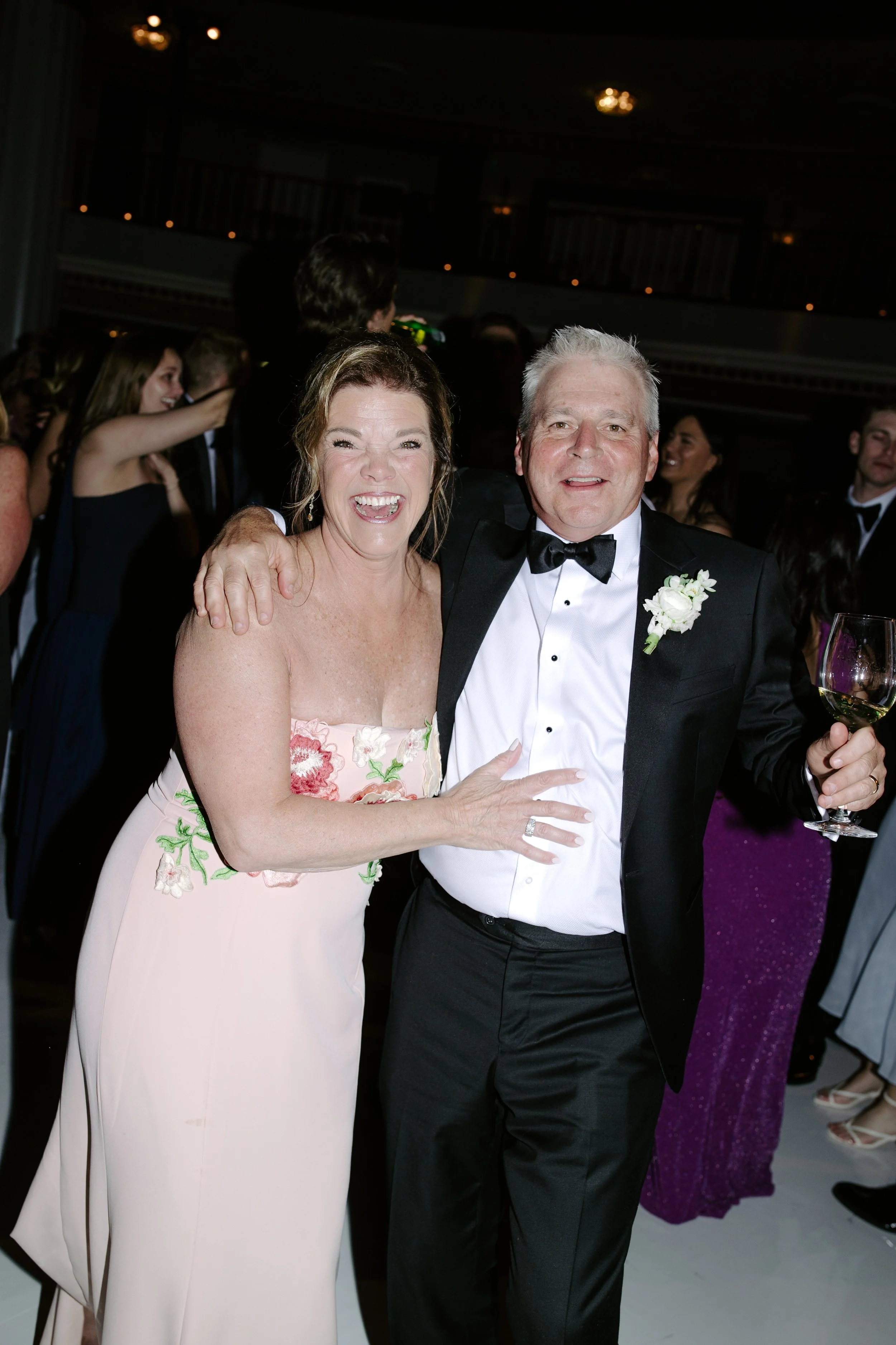 A woman in a floral pink dress and a man in a tuxedo smiling and posing together at a formal event, with other people in the background.
