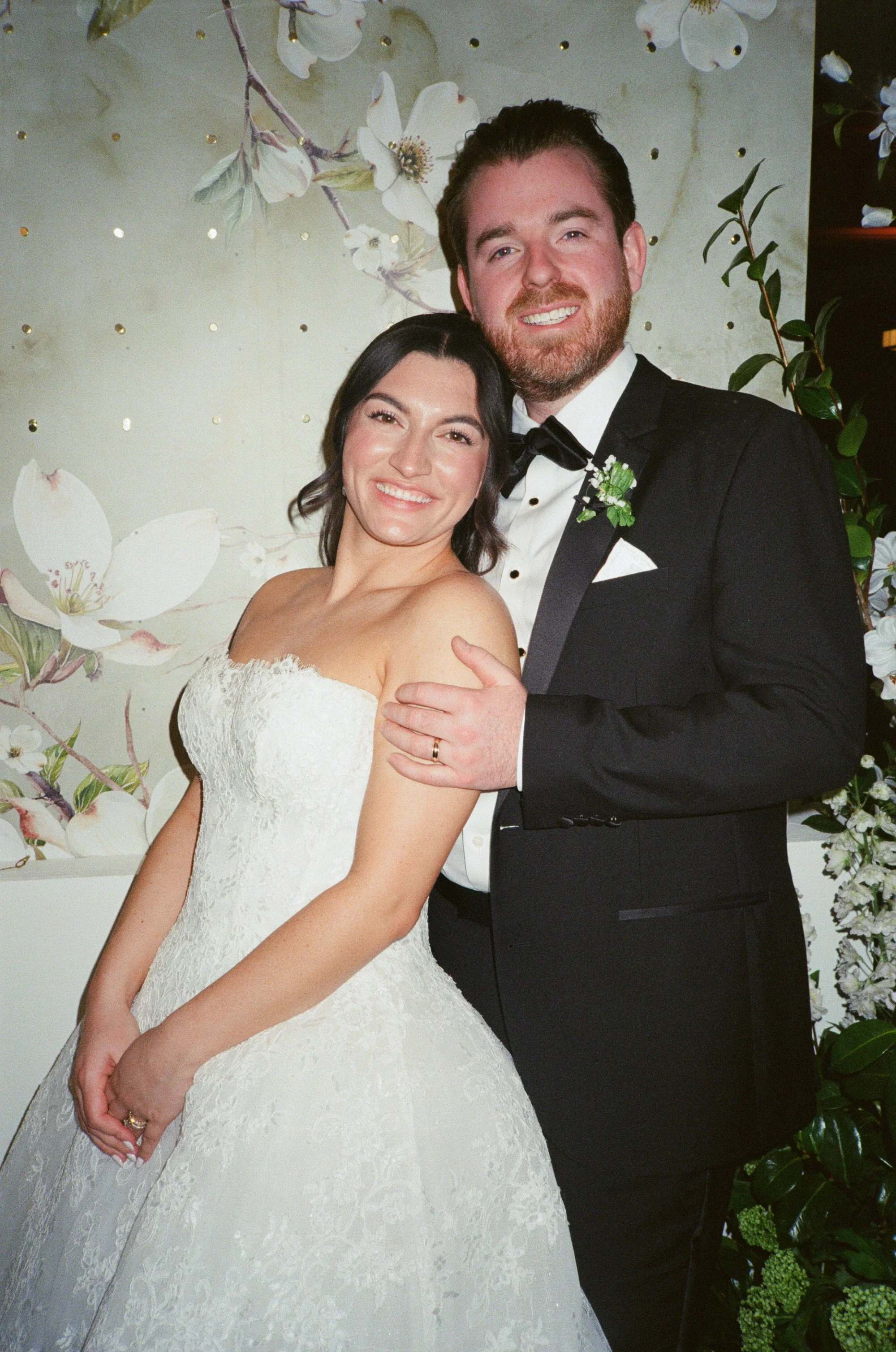 A bride and groom smiling and embracing at their wedding, with a backdrop of white flowers and greenery.