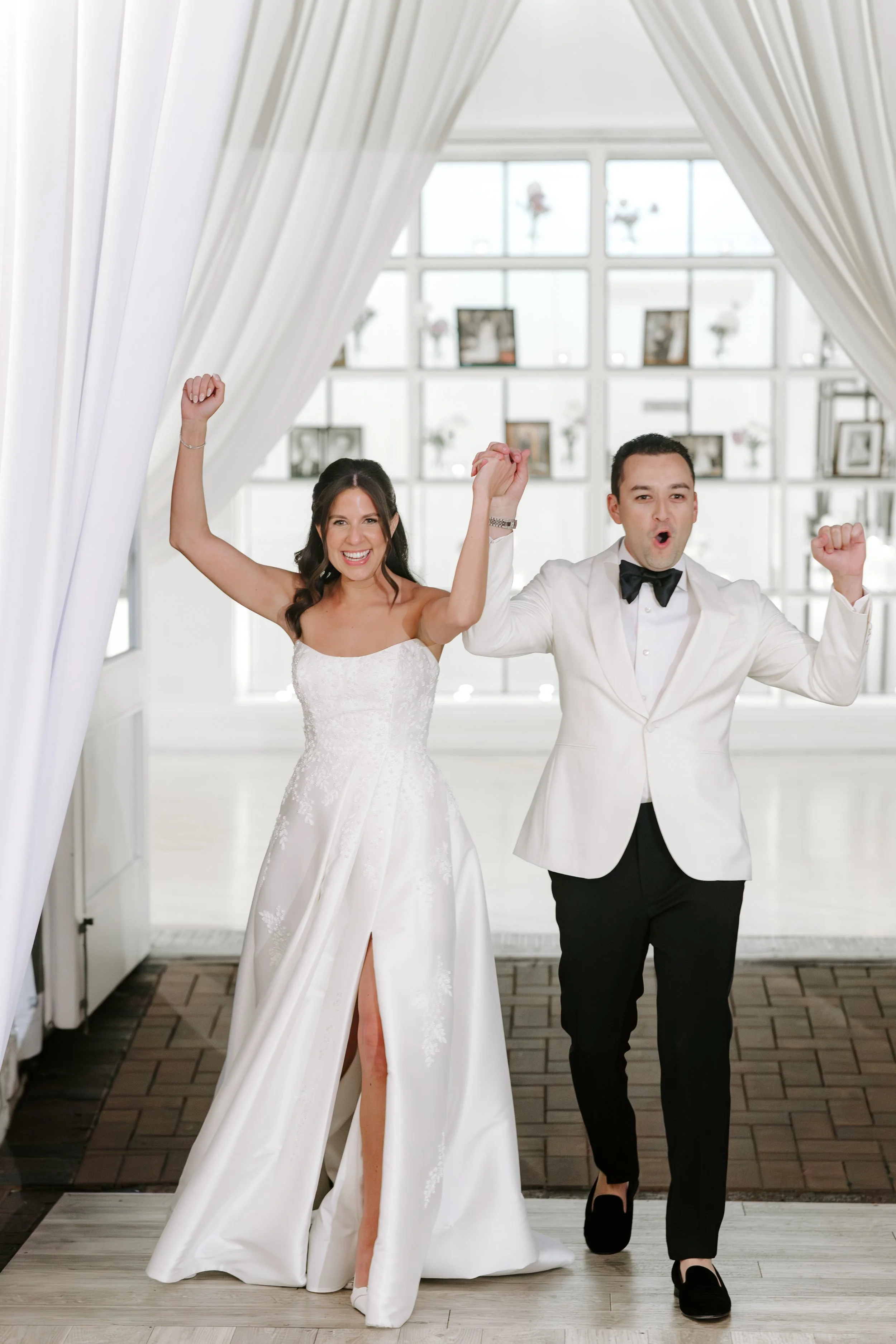 A smiling bride and groom celebrating on their wedding day, with their fists raised in excitement inside a bright, modern venue with large windows and white curtains.