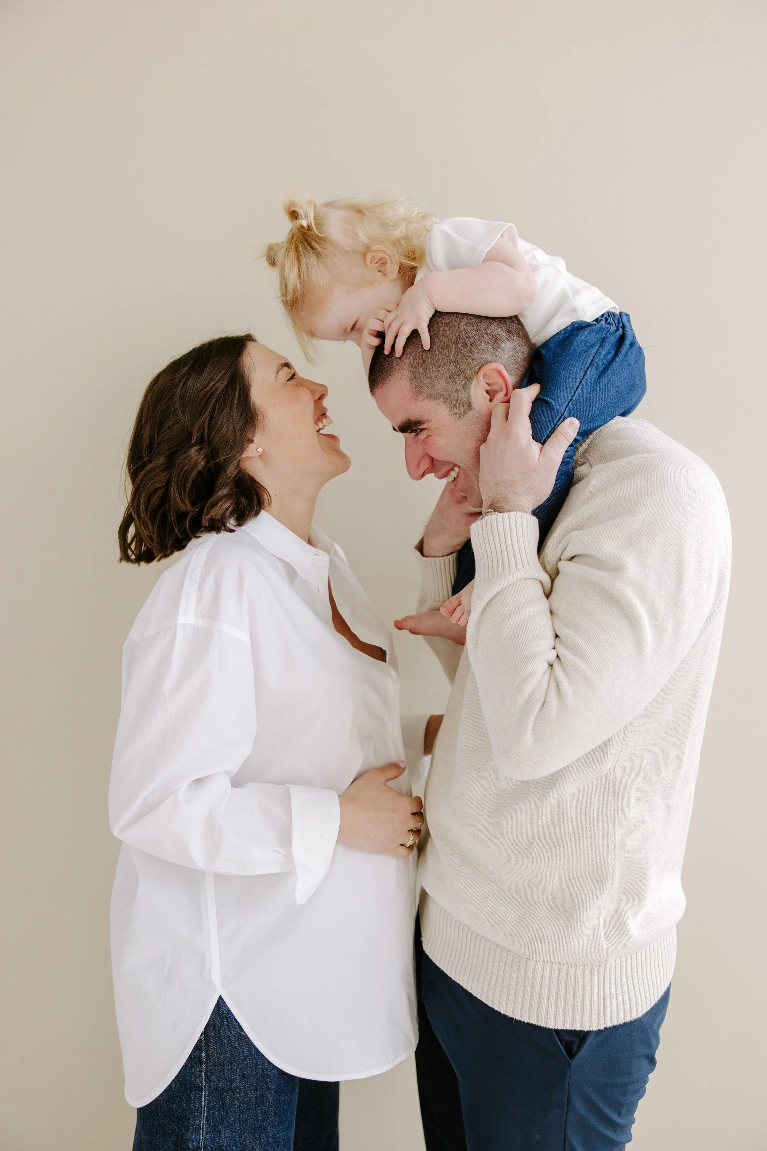 A couple with a pregnant woman and a man, smiling and looking at each other, with a small girl on the man's shoulders, touching his head and leaning in toward him, all sharing a joyful moment.