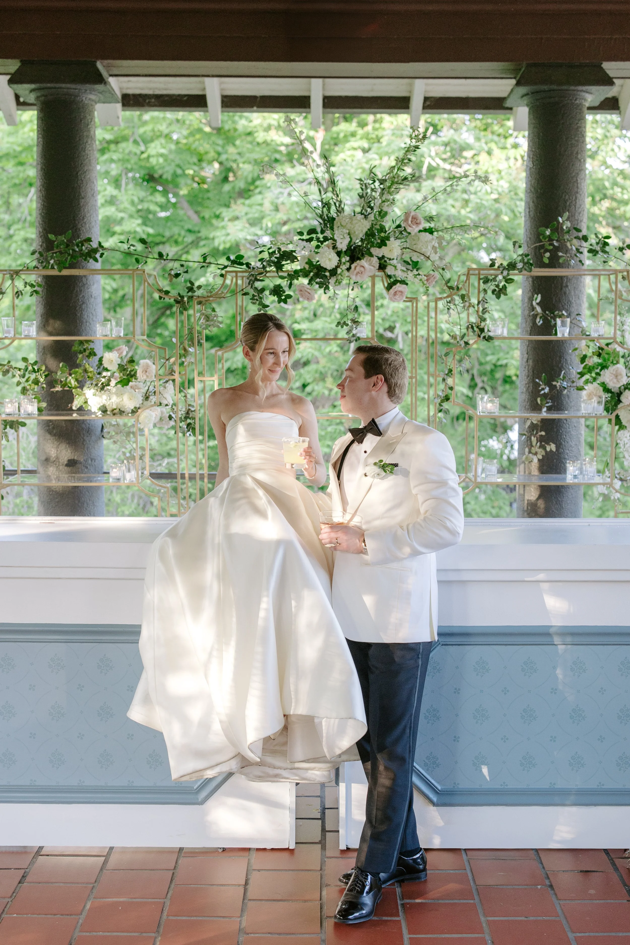 A bride and groom are celebrating at their wedding, standing in front of a decorative floral backdrop with greenery and flowers. The bride is wearing a strapless wedding gown and holding a drink, while the groom is dressed in a white tuxedo jacket wi
