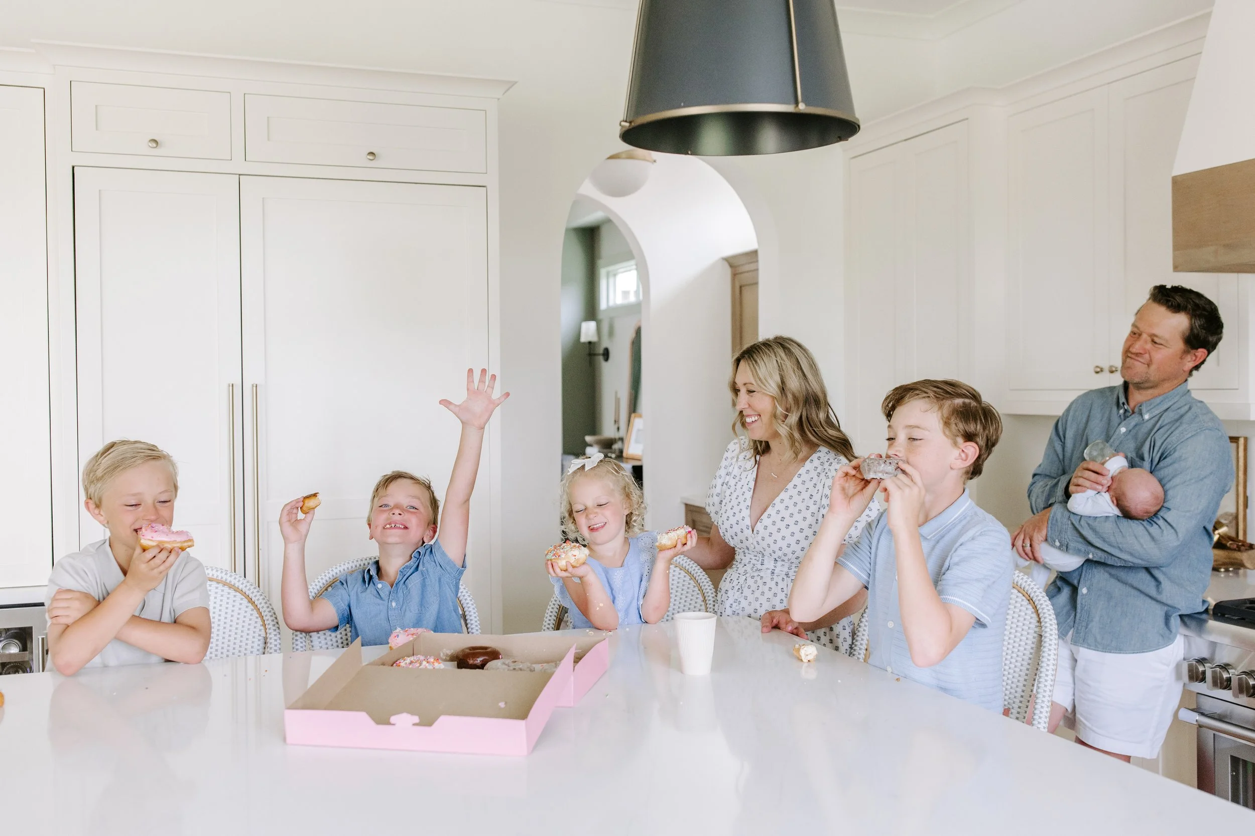 A group of children and adults celebrating a birthday in a bright kitchen with white cabinets, enjoying cake and ice cream at a table, with a pink cake box in the foreground.