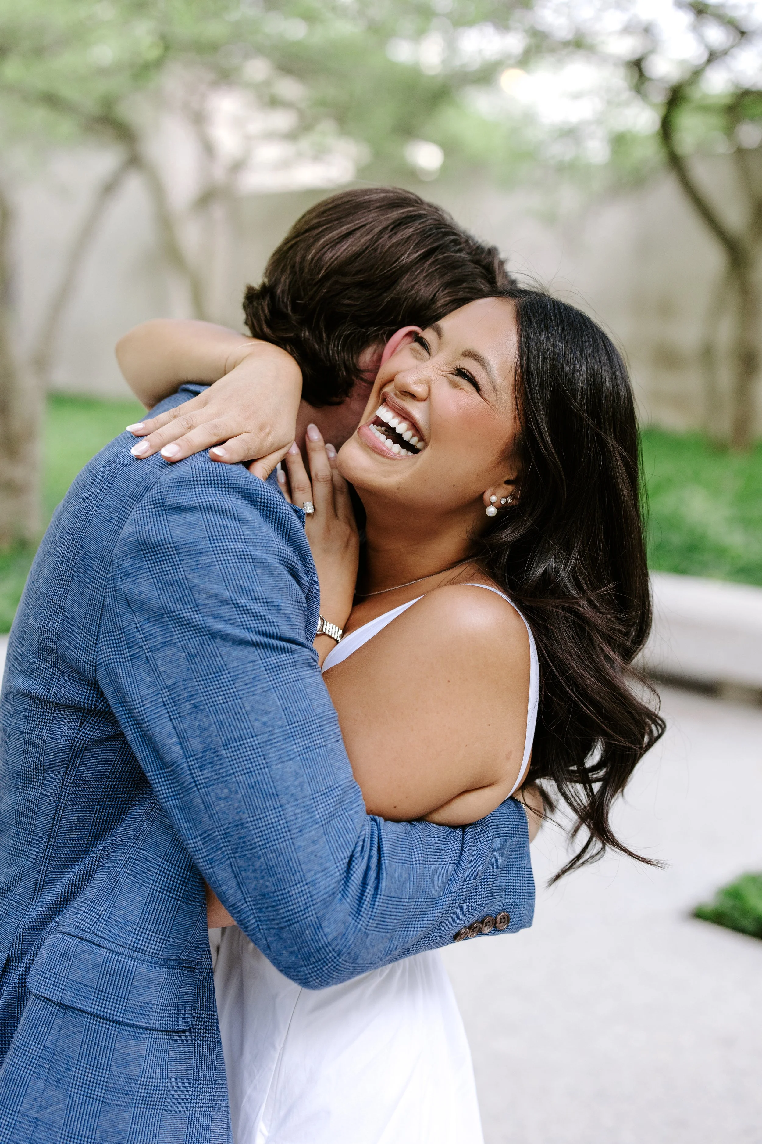 A man with brown hair hugging a woman with black hair outdoors, both smiling joyfully.