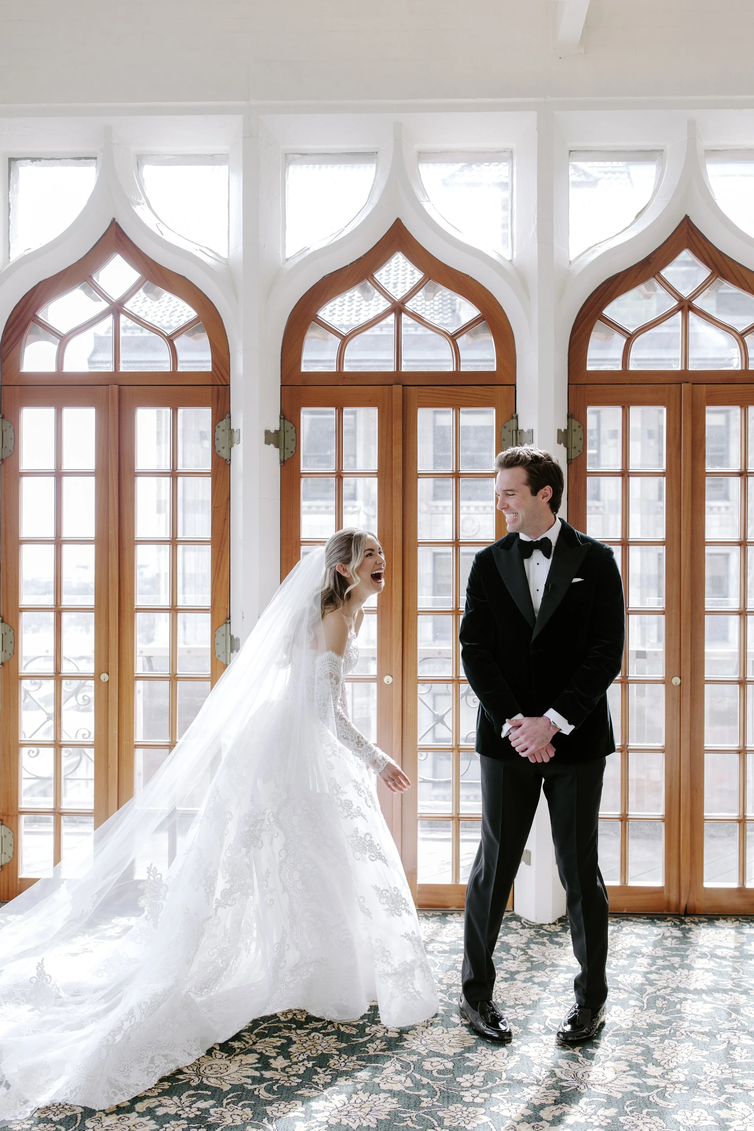 A bride and groom smiling and laughing inside a room with tall wooden doors and large windows. The bride is wearing a white wedding gown with lace details and a long veil. The groom is wearing a black tuxedo with a bow tie. Sunlight streams through t