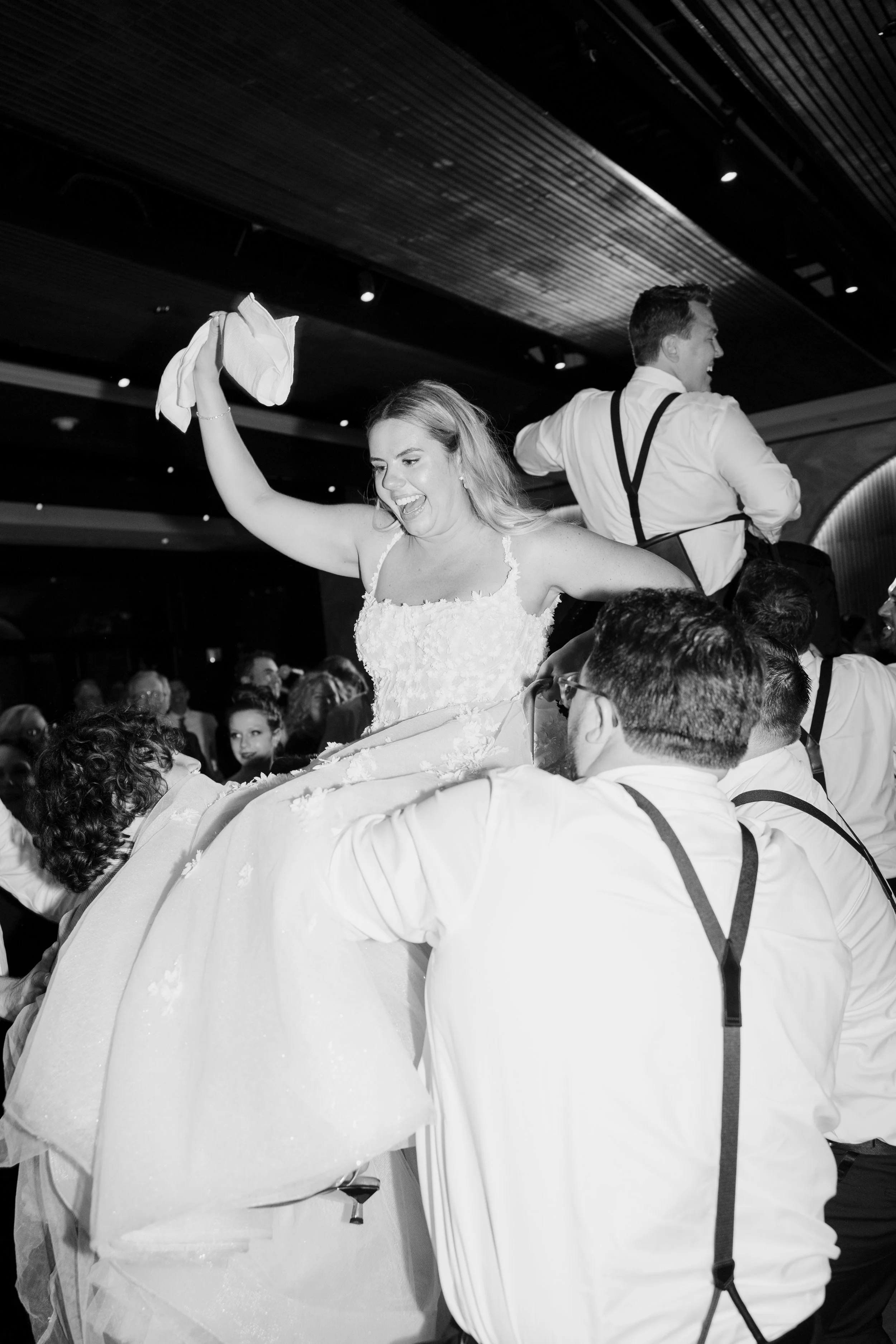A woman in a wedding dress being lifted by multiple men in white shirts with suspenders at a wedding reception.