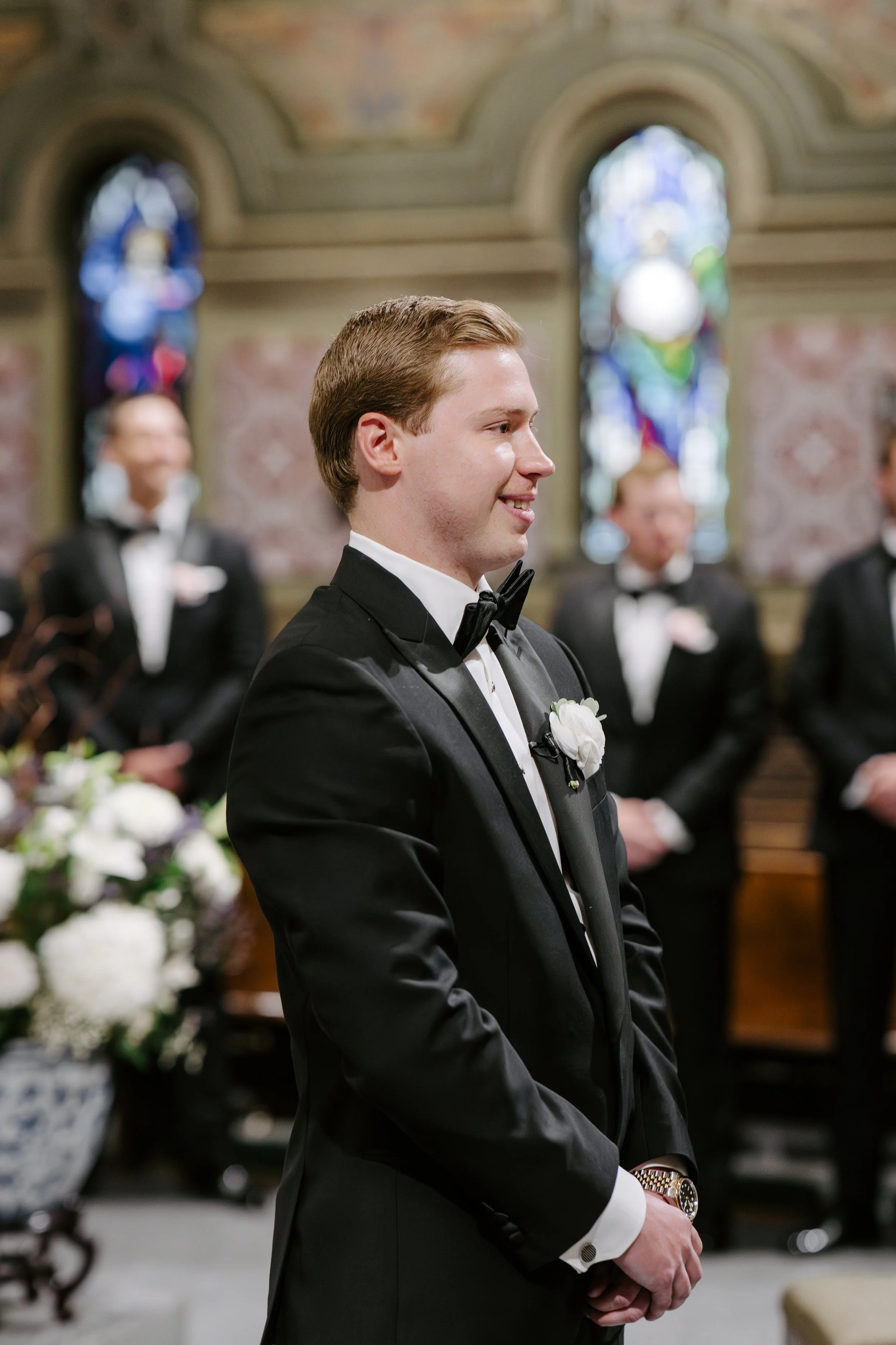 Young man in a tuxedo attending a wedding ceremony in a church, standing with hands clasped in front of him, smiling softly, with stained glass windows in the background.