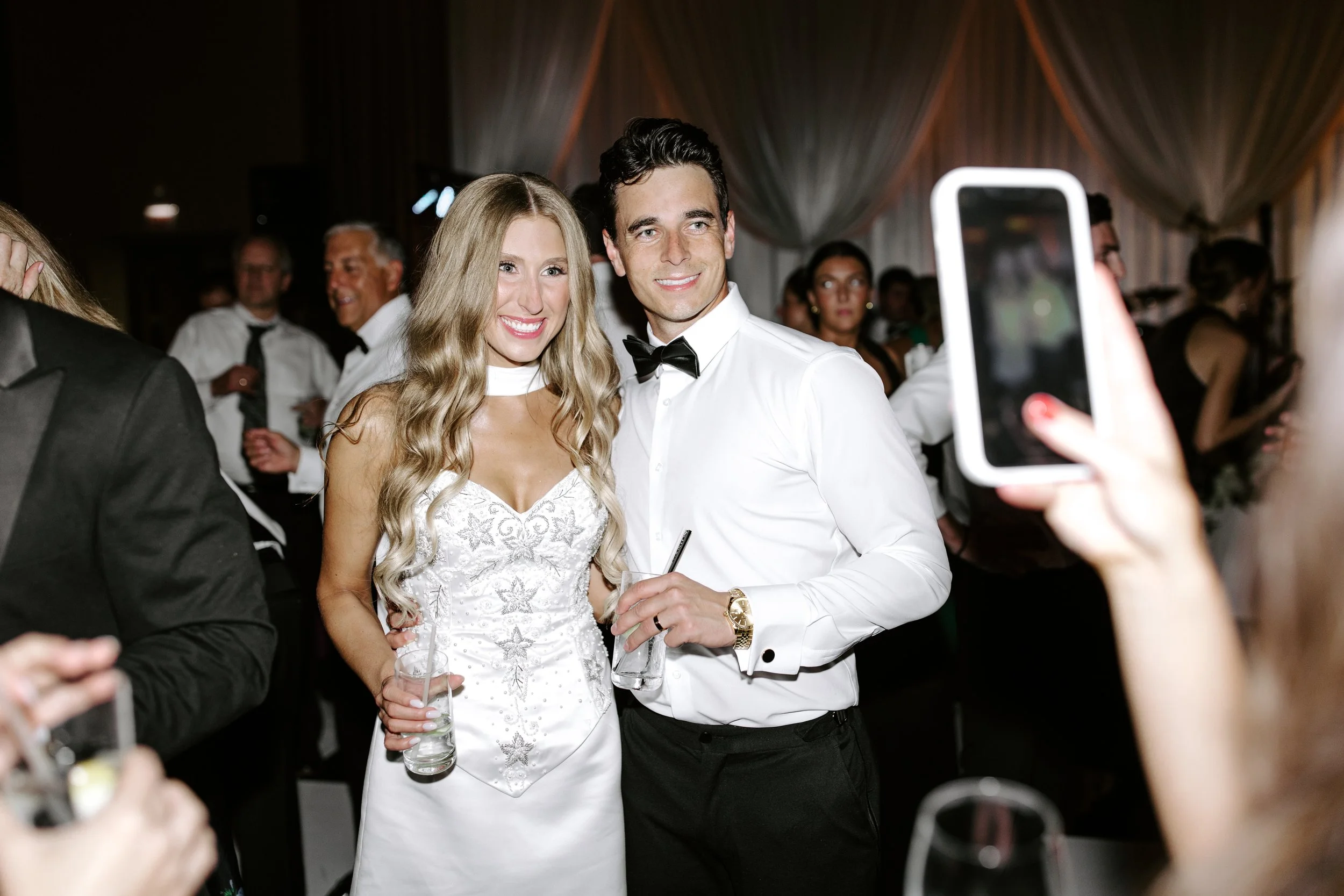 A bride and groom at a wedding reception taking a selfie together, surrounded by guests.