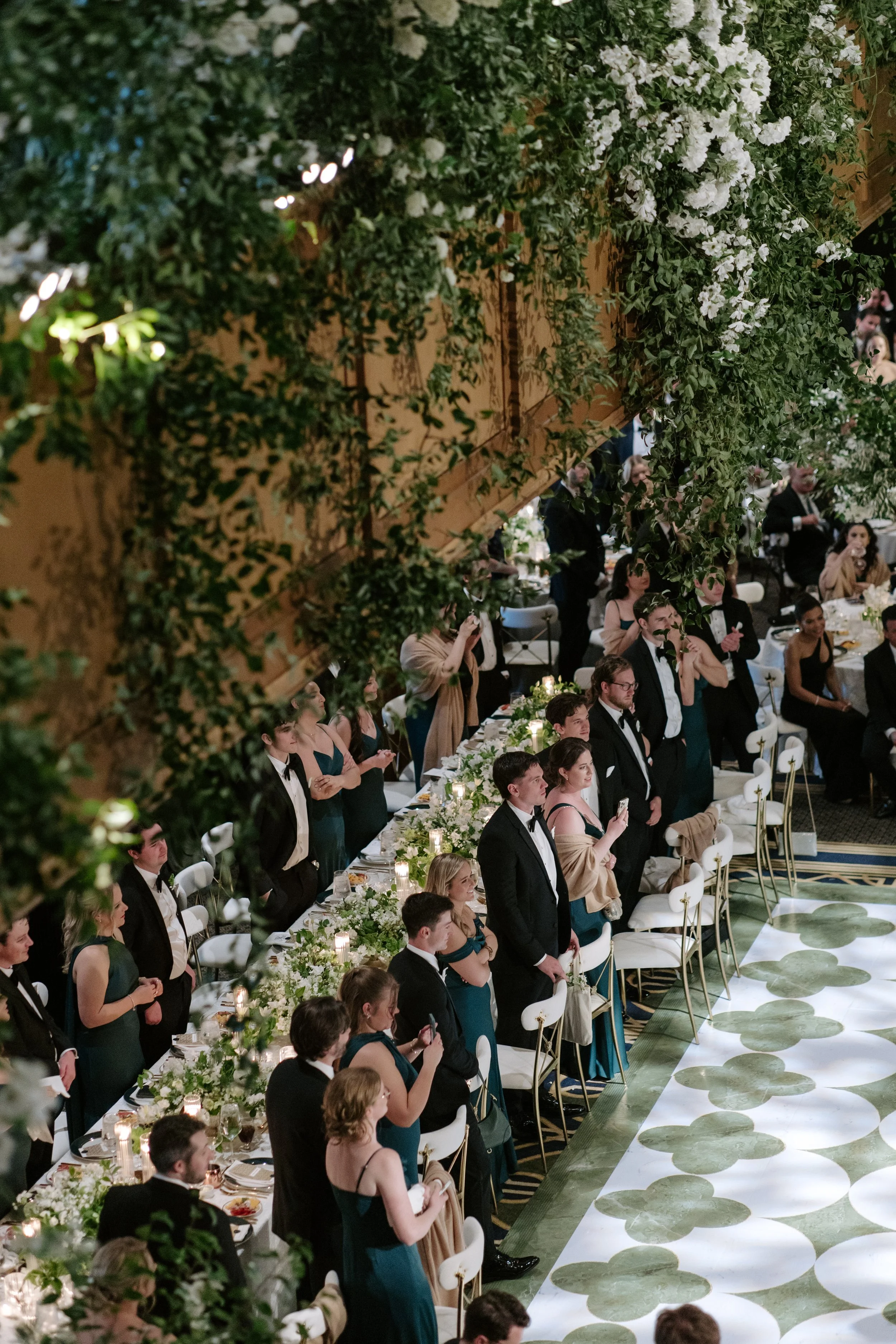 Elegant wedding reception with guests standing and sitting at decorated tables, floral centerpieces, and a green and white patterned aisle runner, surrounded by lush greenery and flowers.