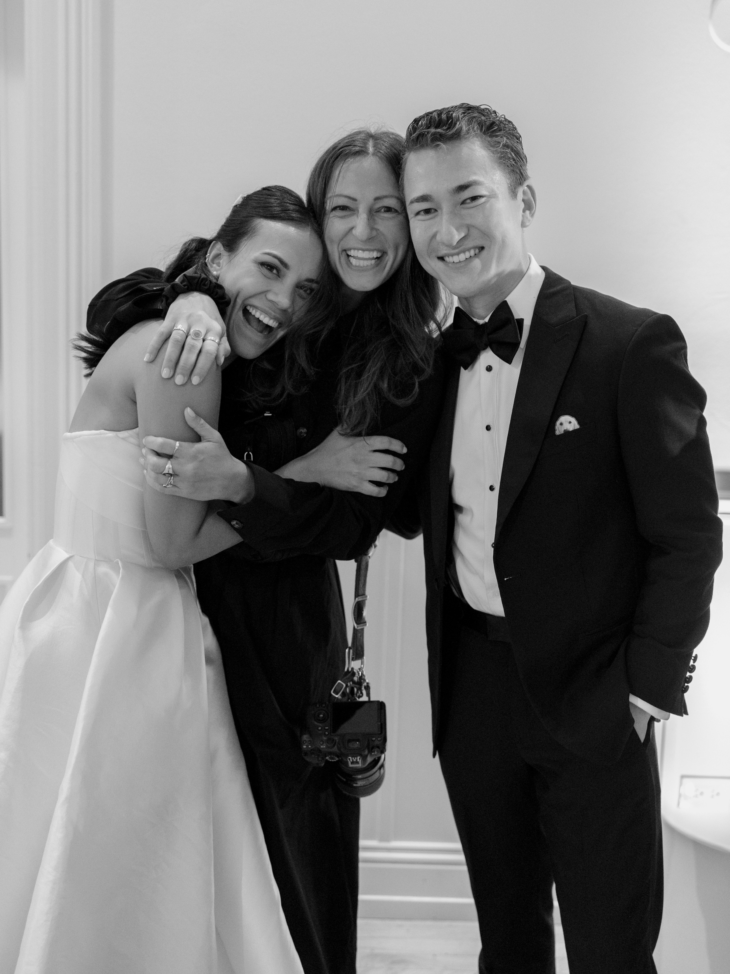 A group of three people smiling and hugging each other at an indoor event, one holding a camera, dressed in formal attire including tuxedo and dress.