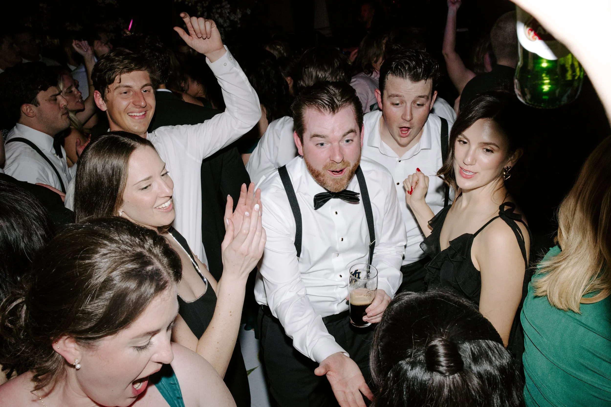 People dancing and socializing at a lively party, dressed in formal and casual attire, with drinks in hand, in a crowded indoor setting.