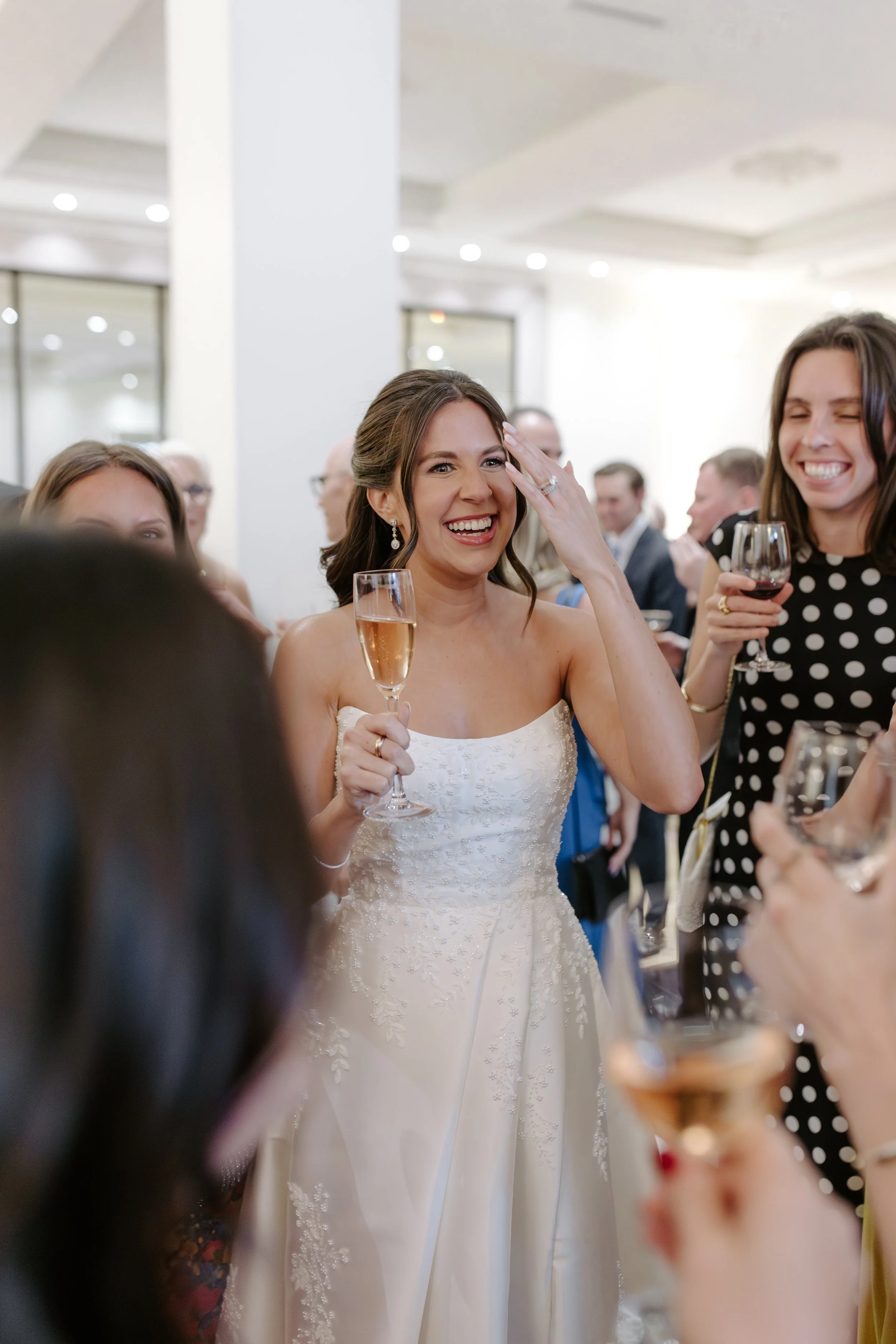 A woman in a wedding dress smiling and holding a glass of champagne at a celebration, surrounded by guests in formal attire.