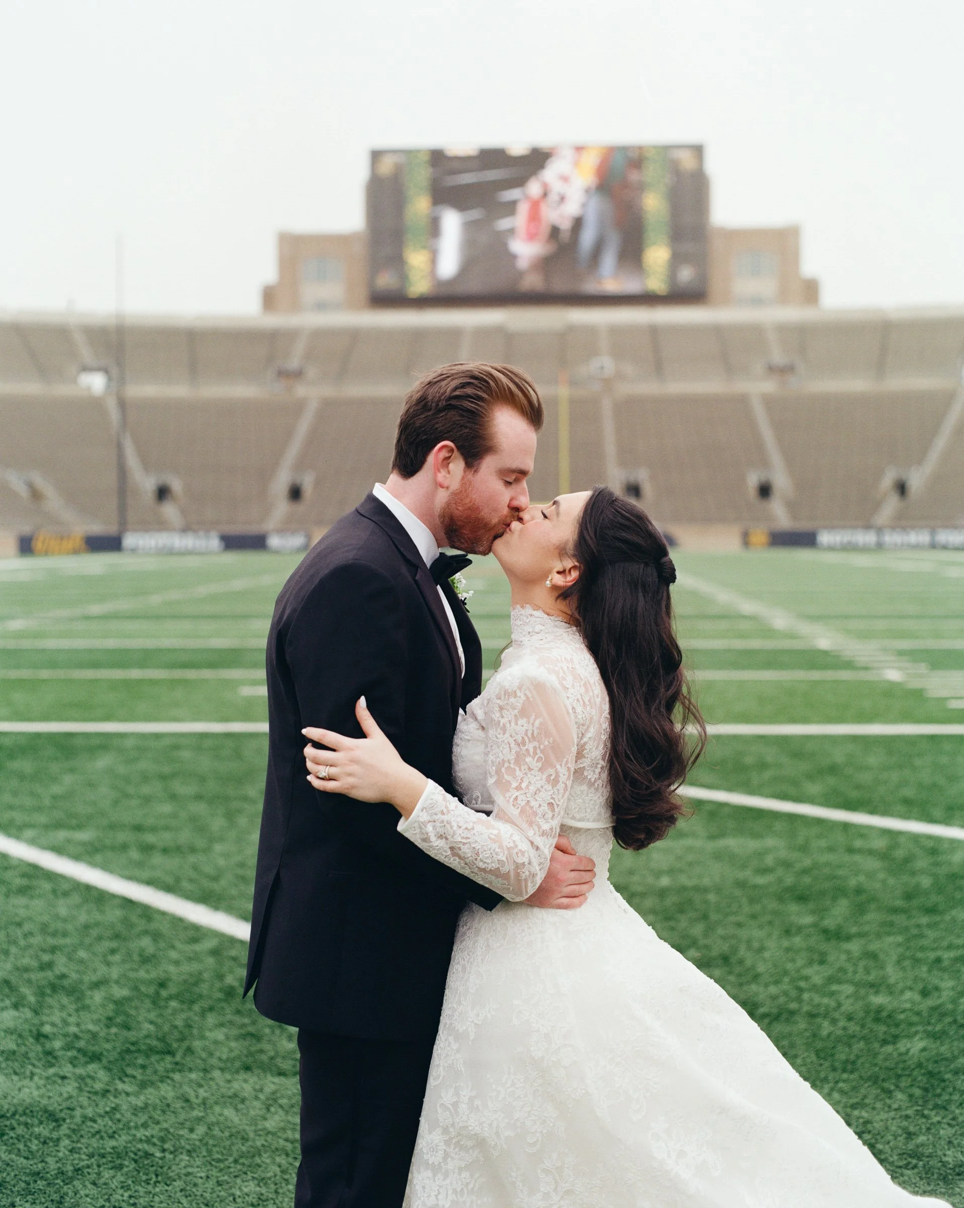 A bride and groom sharing a kiss on a football field at a stadium, with a large scoreboard behind them showing a blurred image of other people.