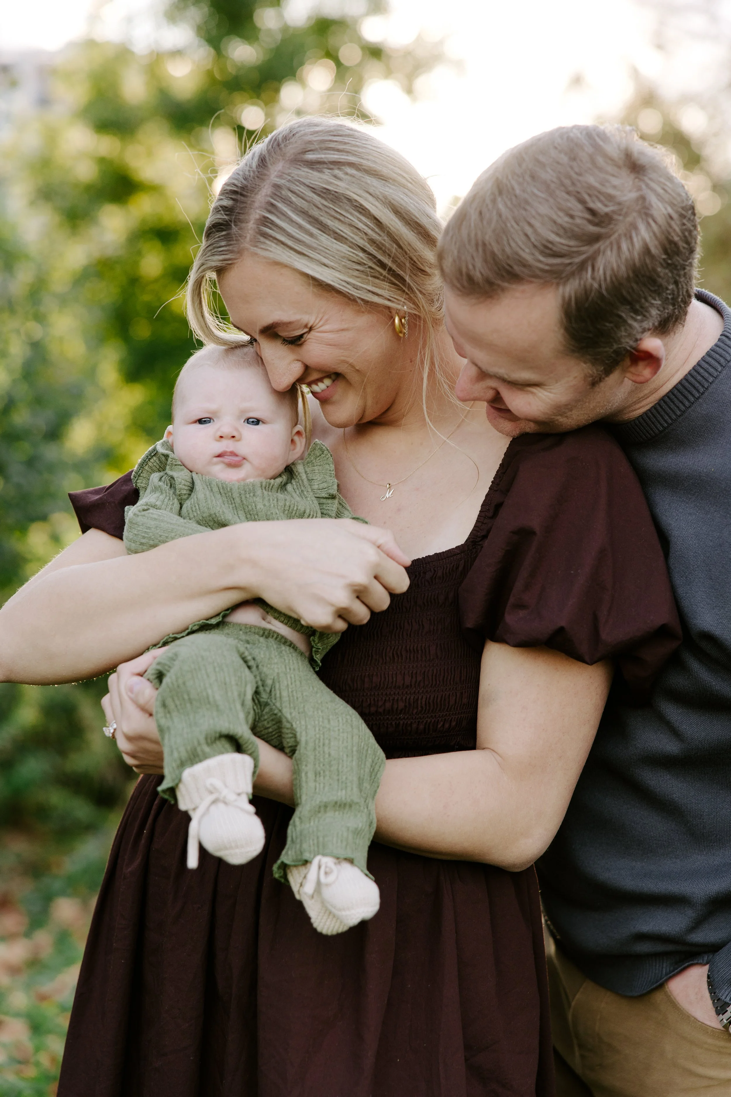 Close-up of a family of three outdoors in a park, with a woman holding a baby while a man leans in close, all smiling and happy.