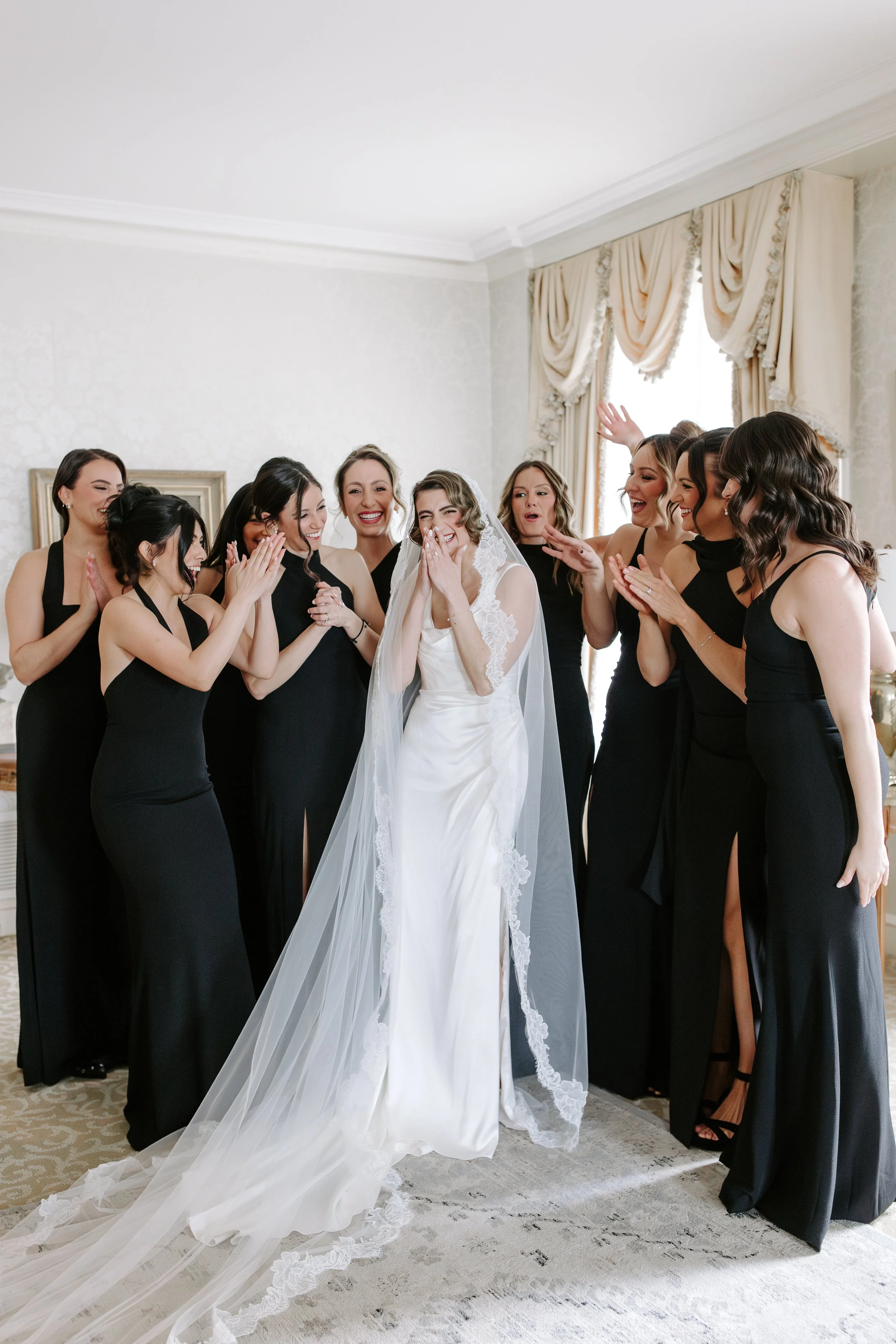 A bride in a white wedding dress surrounded by eight women in black dresses, all smiling and celebrating indoors near a window with cream-colored curtains.