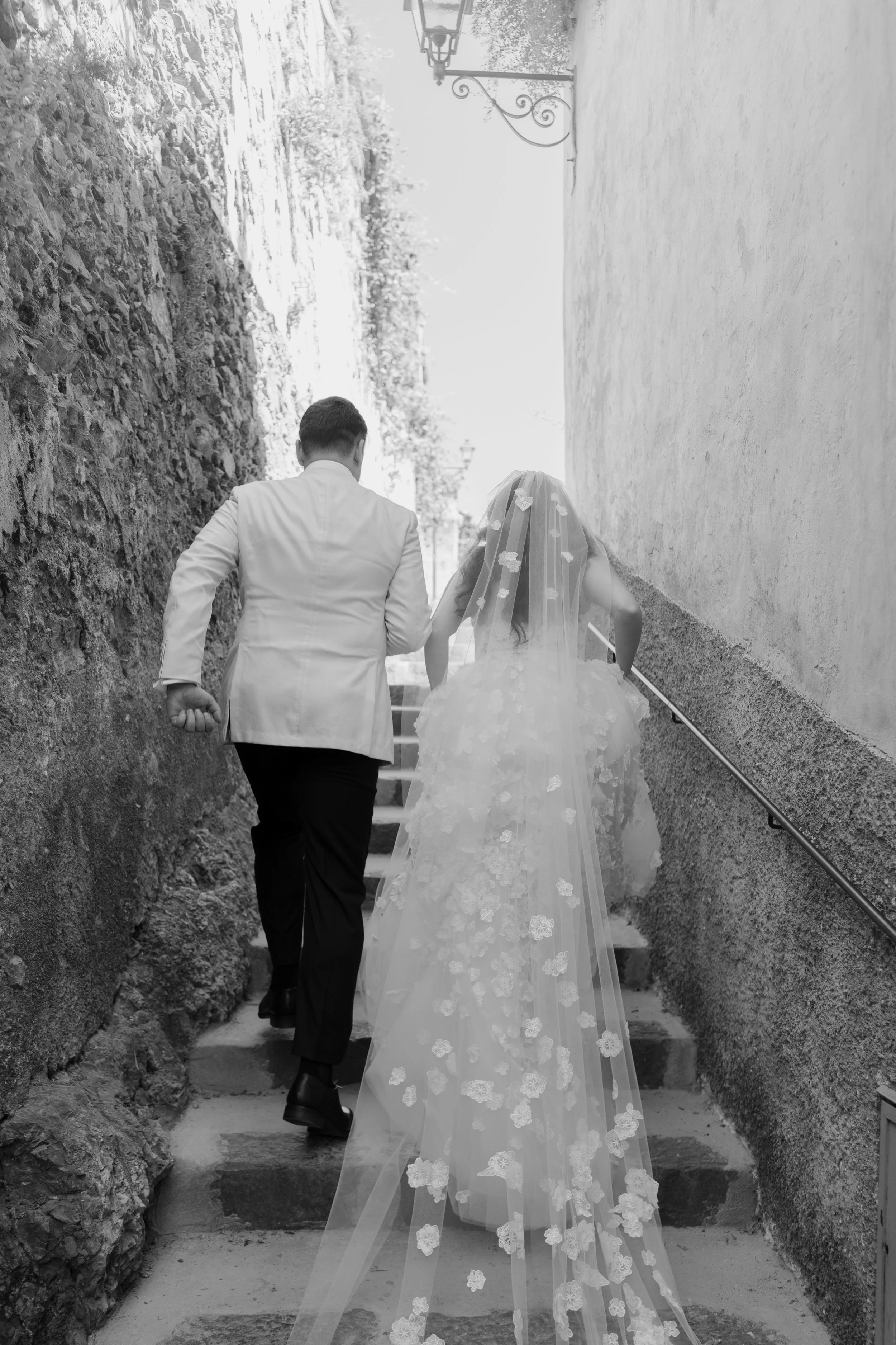A groom and a bride walking up stairs outdoors; the bride wears a long wedding gown with a veil covered in floral appliqués, and the groom is dressed in a white tuxedo jacket and black pants.