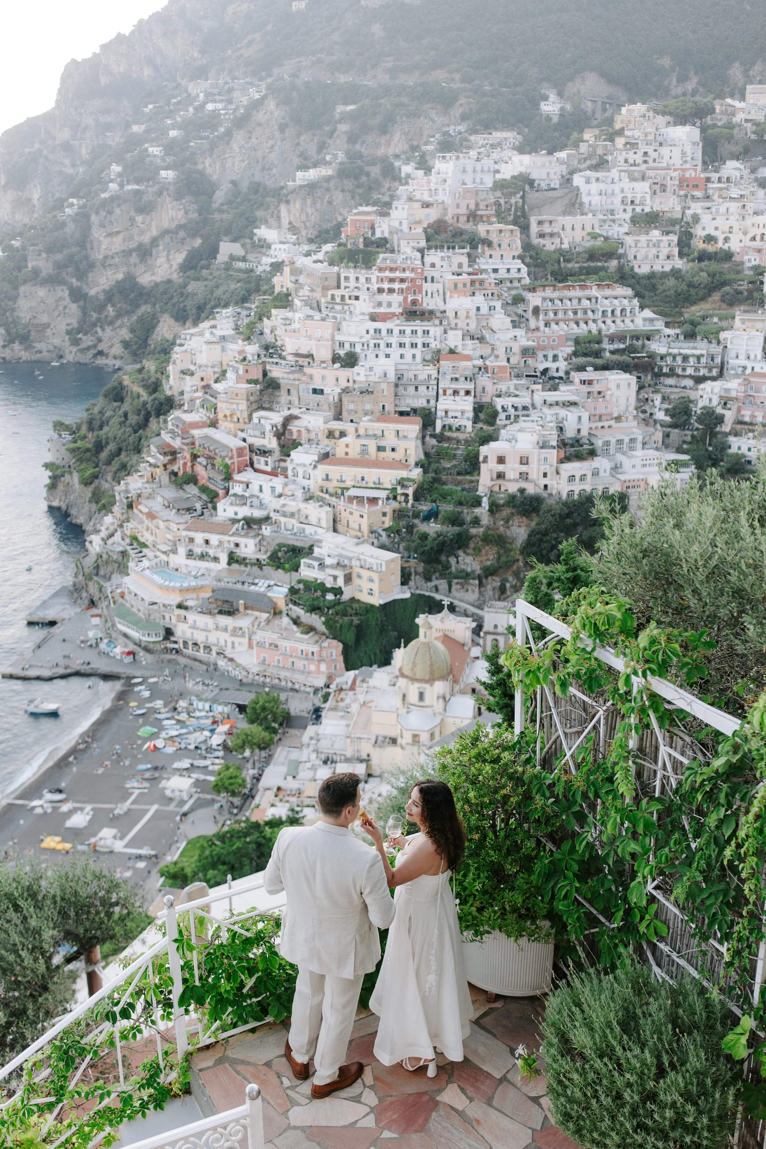 A couple dressed in white, standing on a balcony overlooking a coastal town with white and pastel-colored buildings on a hillside and a body of water with boats, with green plants and trees around them.