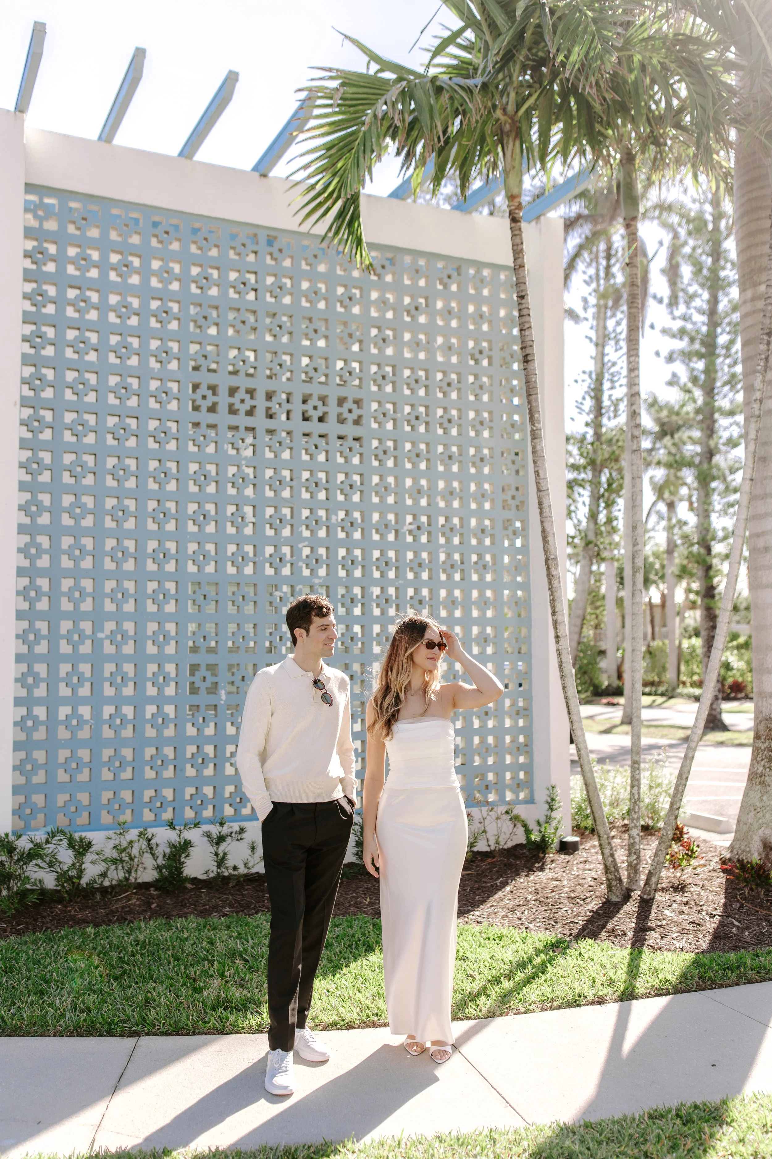A young man and woman standing outdoors next to a decorative white wall with geometric cutouts, surrounded by palm trees and greenery, on a sunny day.