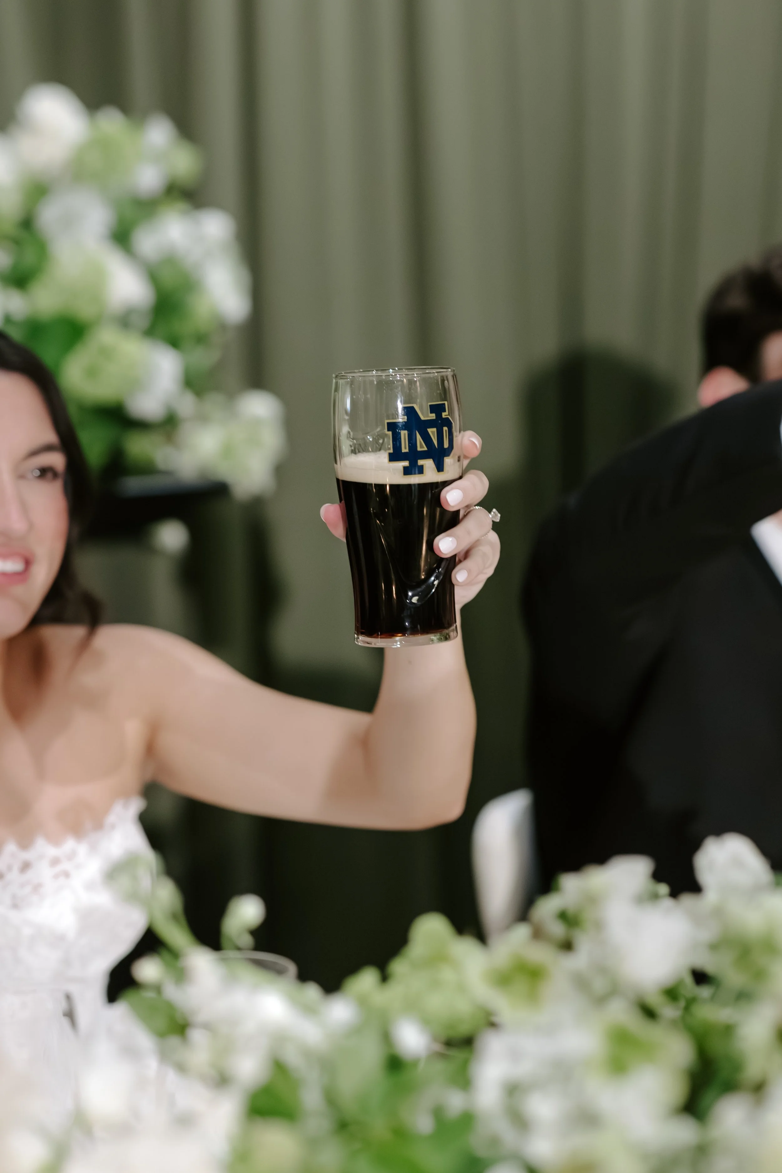 A woman in a wedding dress holding up a dark beer in a glass with a blue and gold number 15 logo, at a celebration or wedding with floral decorations and green curtains in the background.