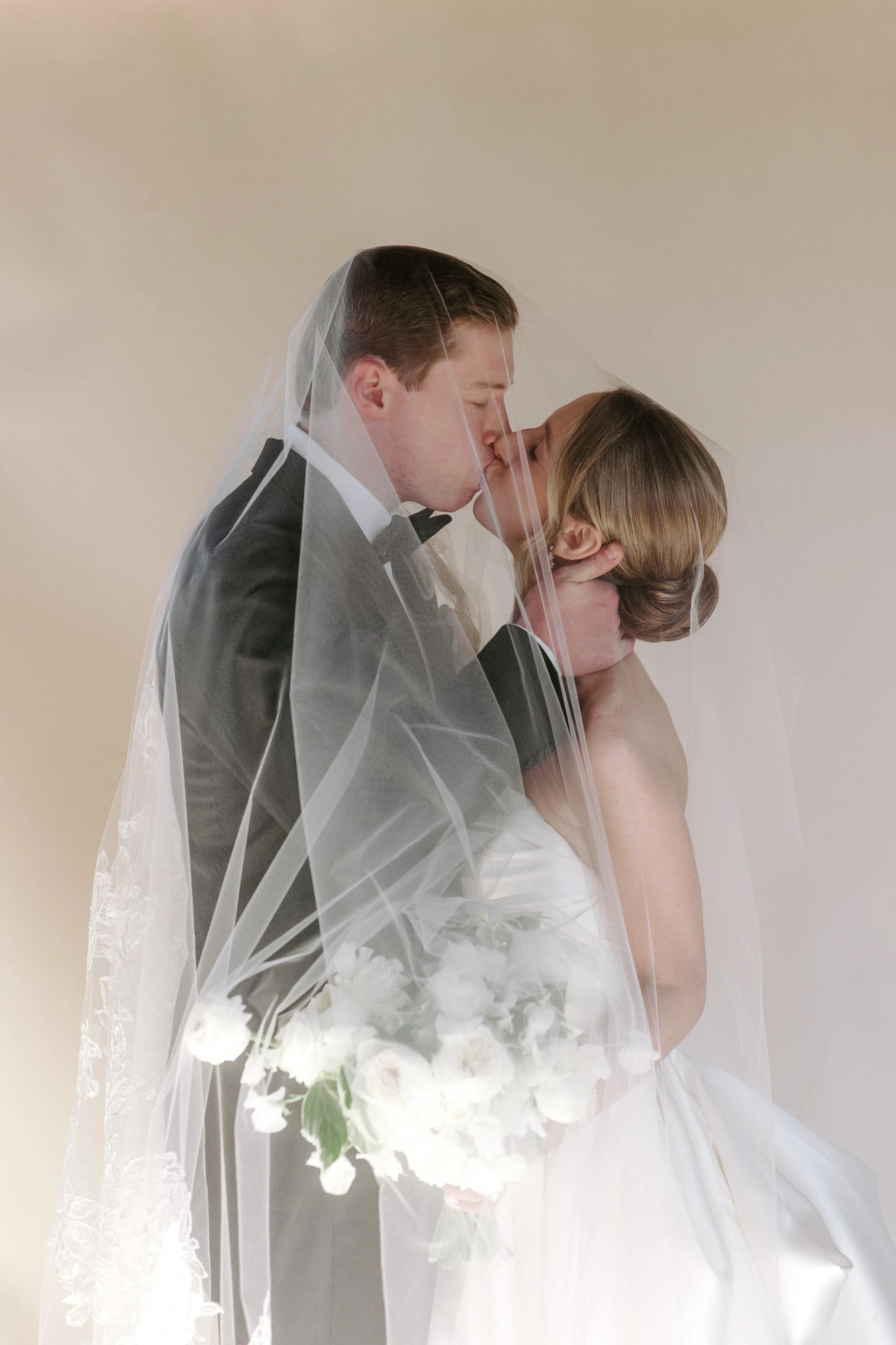 A man and woman, dressed as a bride and groom, share a kiss under a veil, with the woman holding a bouquet of white flowers.