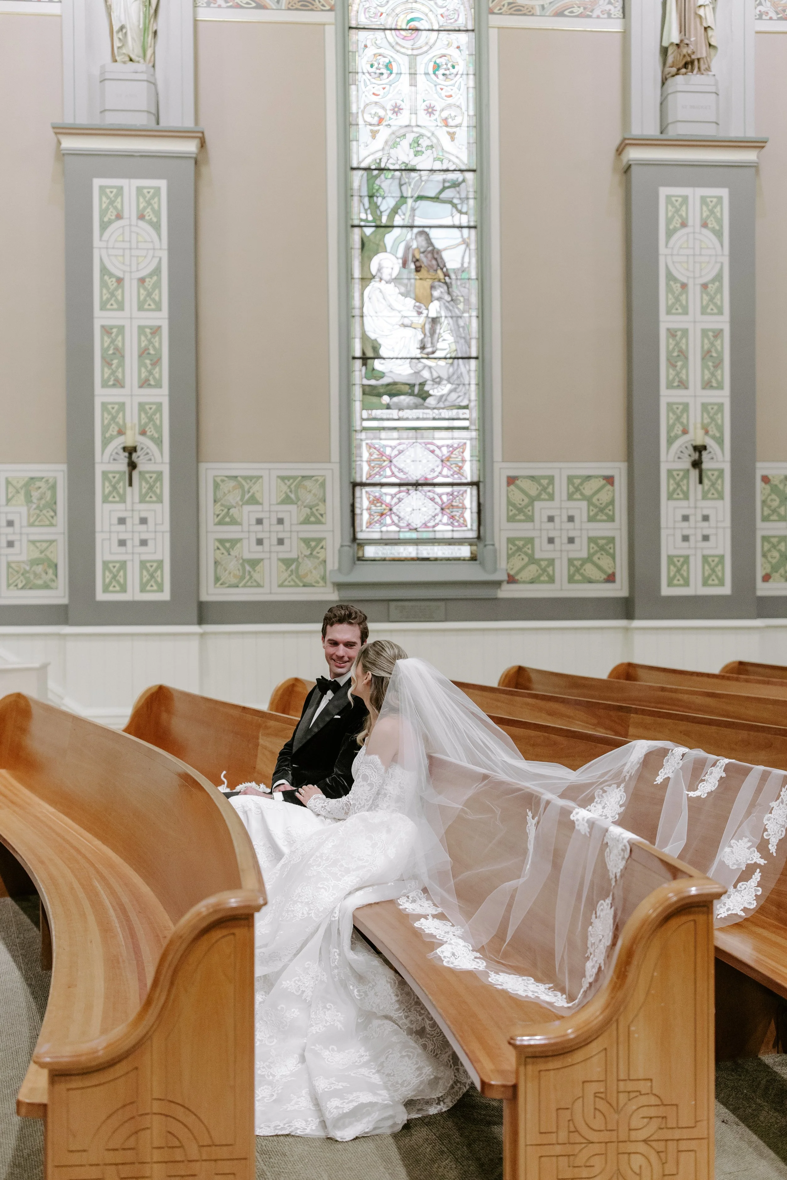A bride and groom sitting in a church pew, smiling at each other, with a stained glass window behind them, and the bride wearing a long white wedding dress with a veil.