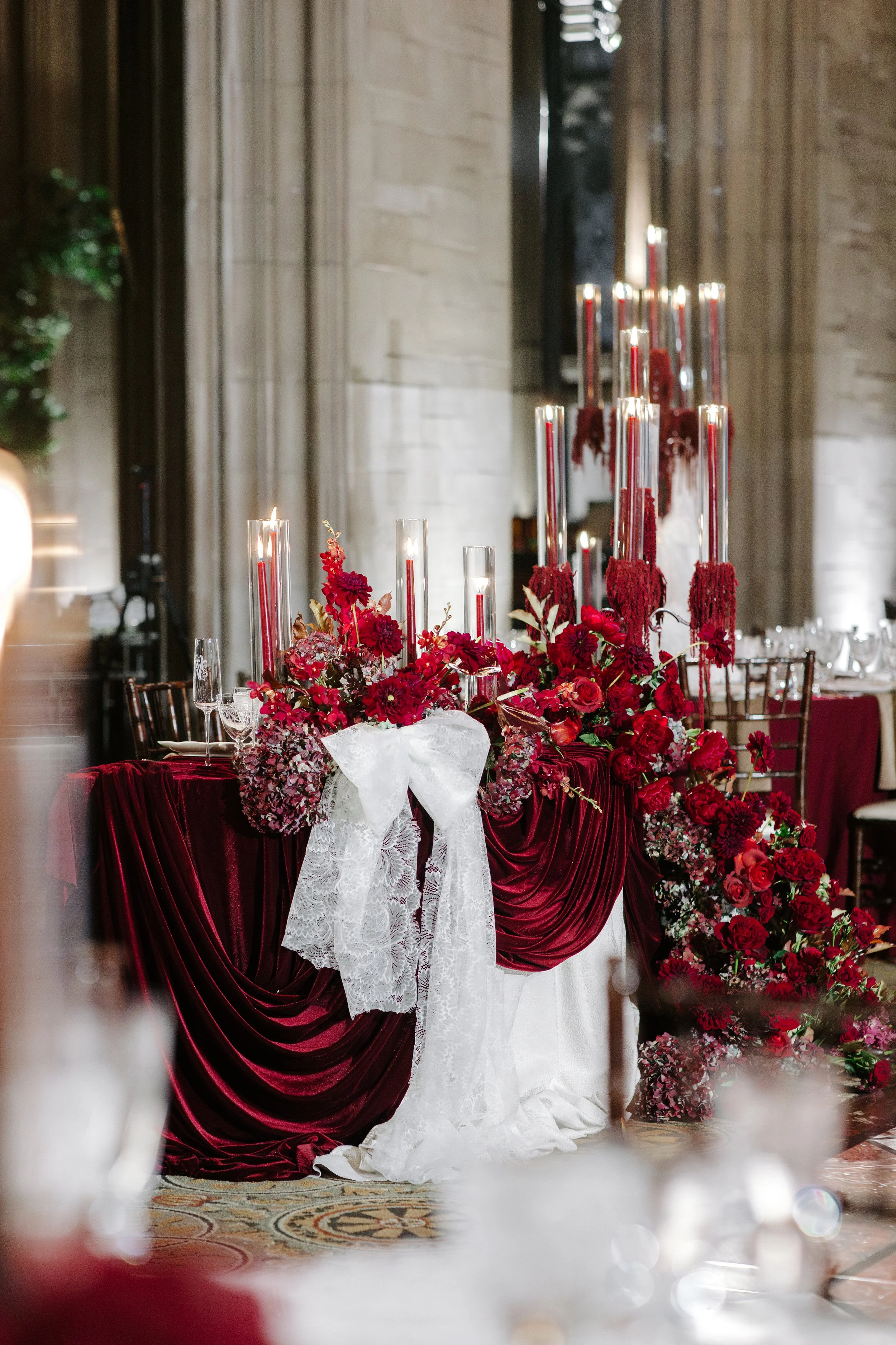 Elegant wedding table decoration featuring red flowers and tall red candles with a white lace bow, set against a church-like backdrop with stone walls.