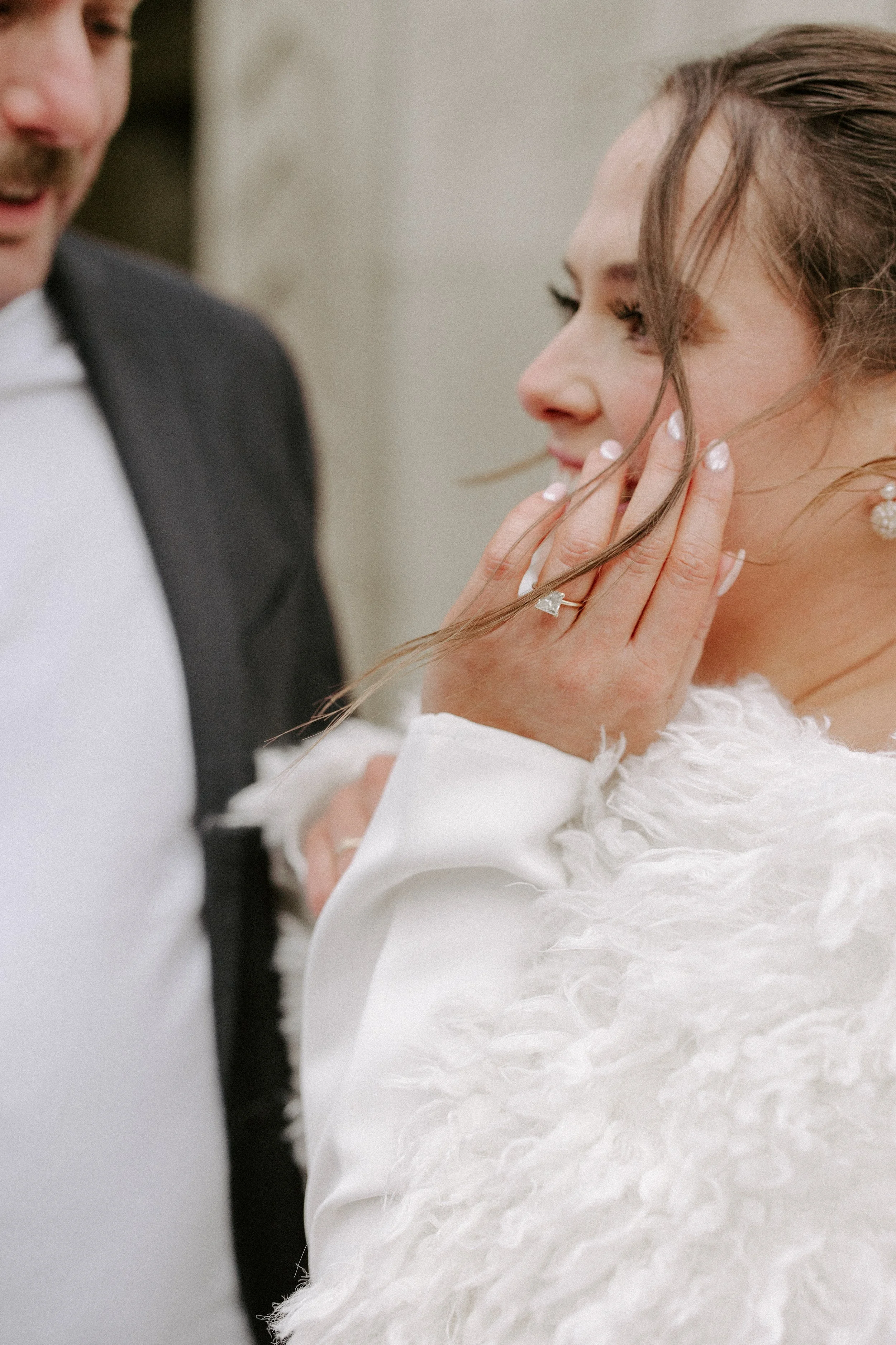 A bride with brown hair in soft waves, wearing pearl earrings and a white wedding dress with textured fabric, looks to the side while holding her face gently.