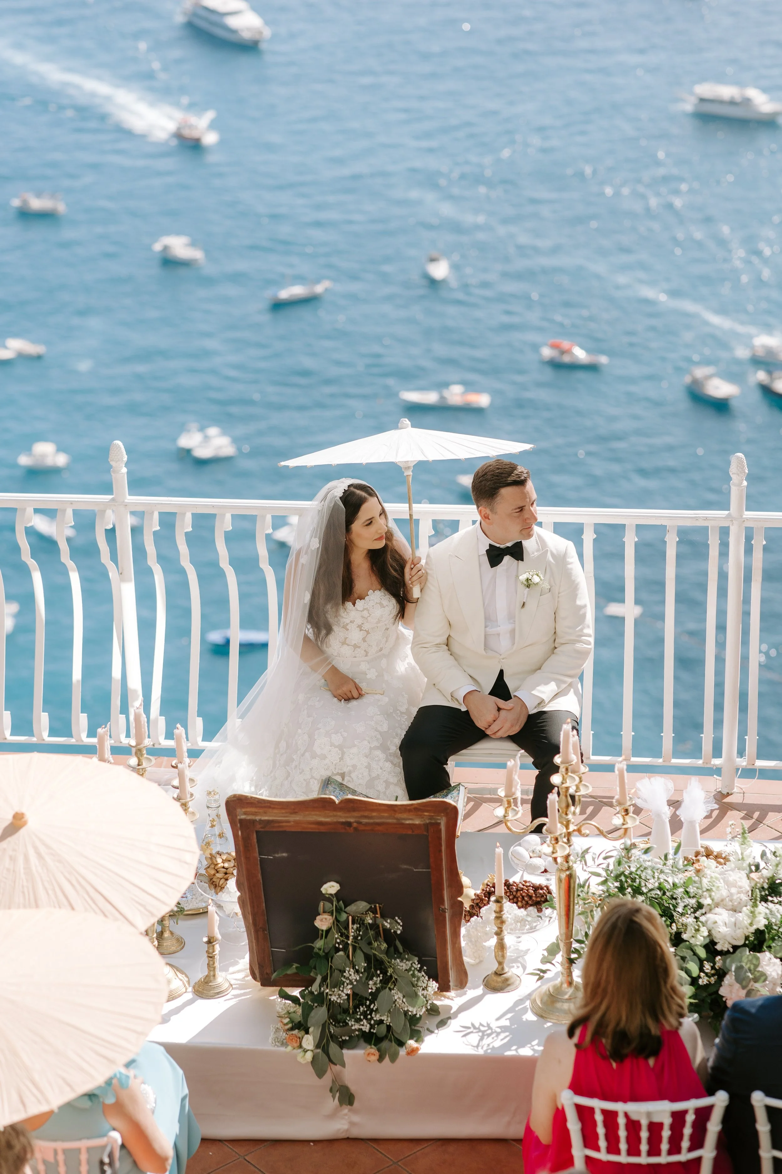 A bride and groom sitting together during their wedding ceremony on a balcony overlooking a body of water with boats. The bride is holding a white parasol and wearing a white wedding dress, while the groom is dressed in a white tuxedo with a black bo