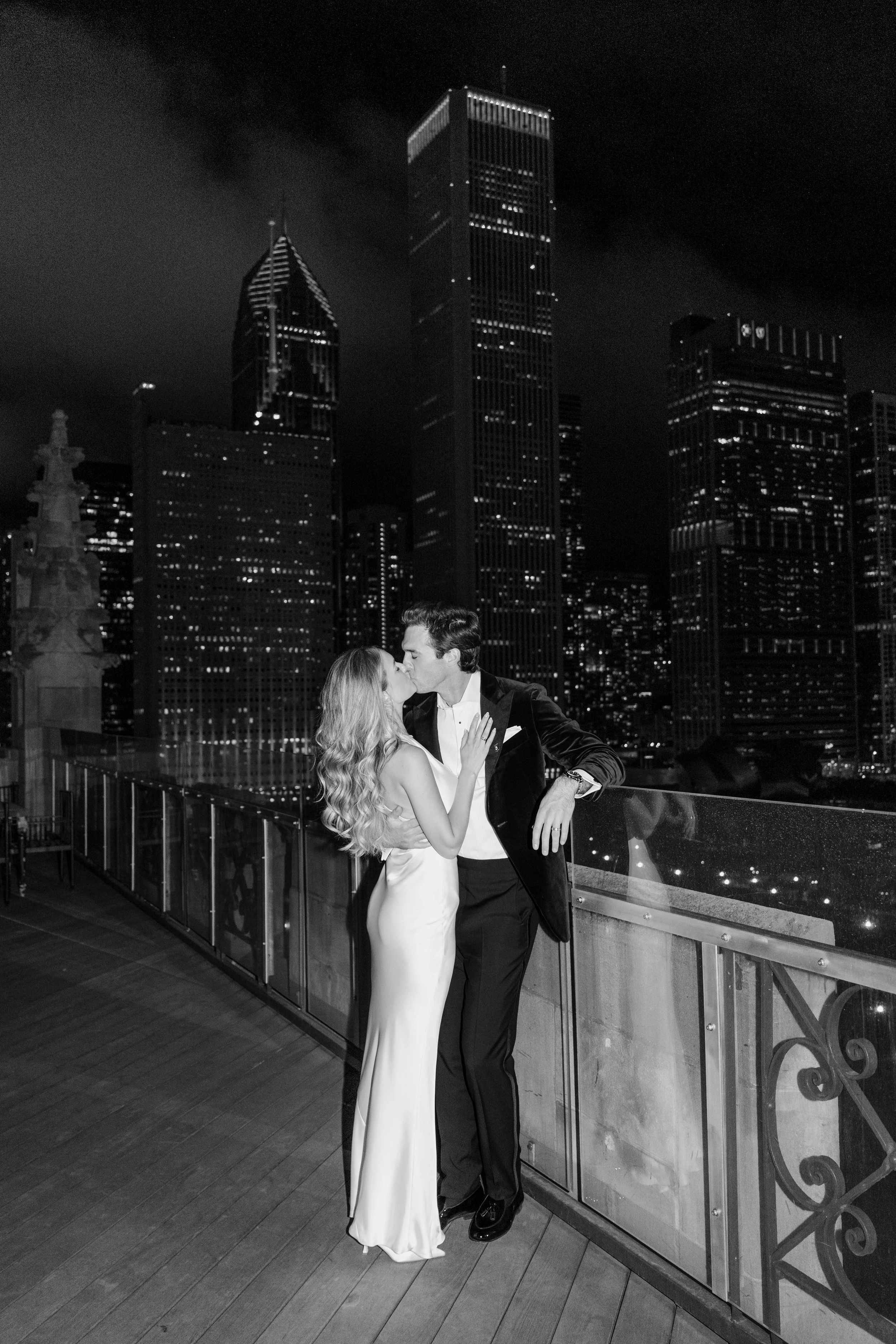 A black-and-white photo of a couple kissing on a rooftop at night with a city skyline featuring tall skyscrapers in the background.