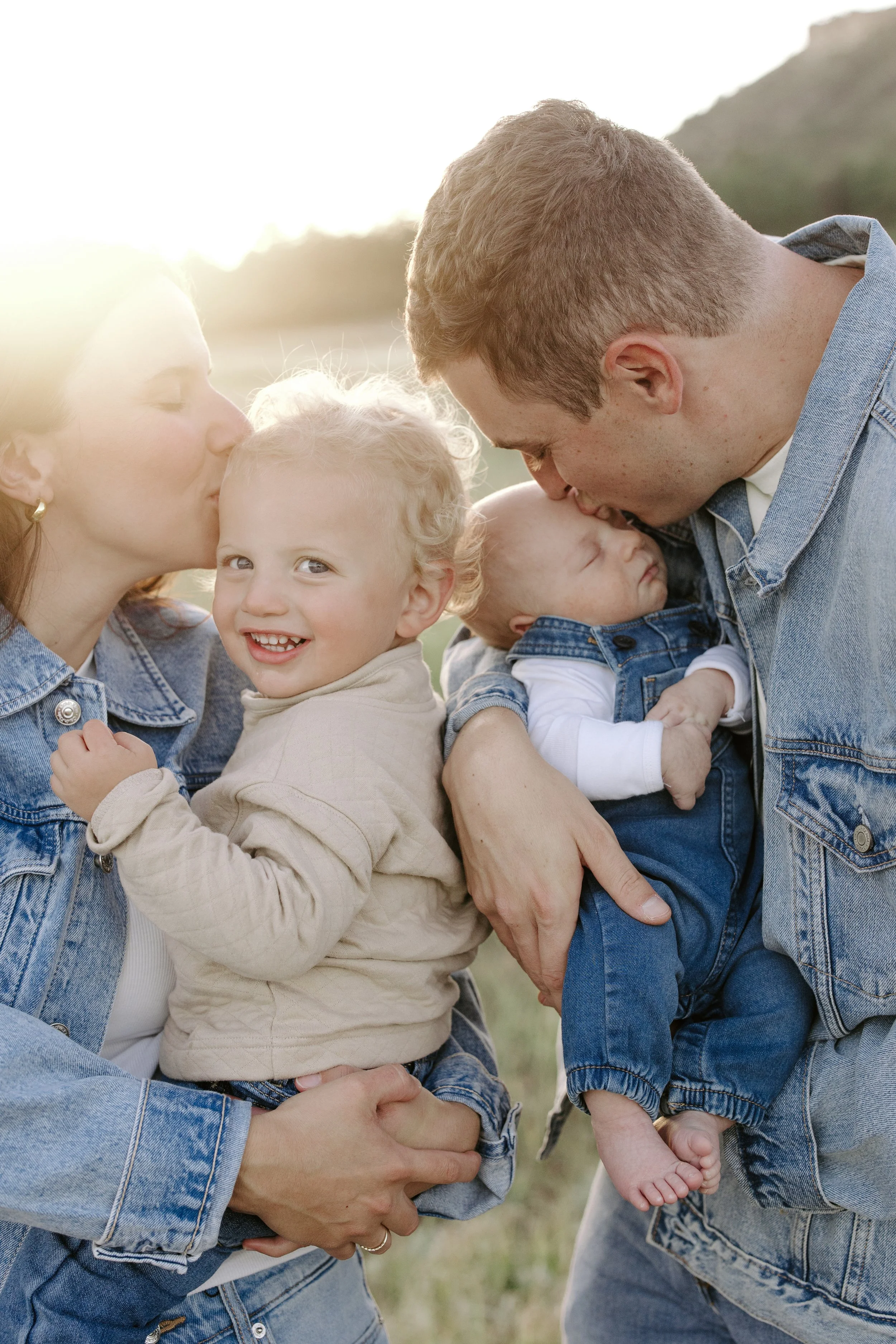 A family of four outside during sunset, featuring a woman, a man, and two young children, all sharing affectionate moments.