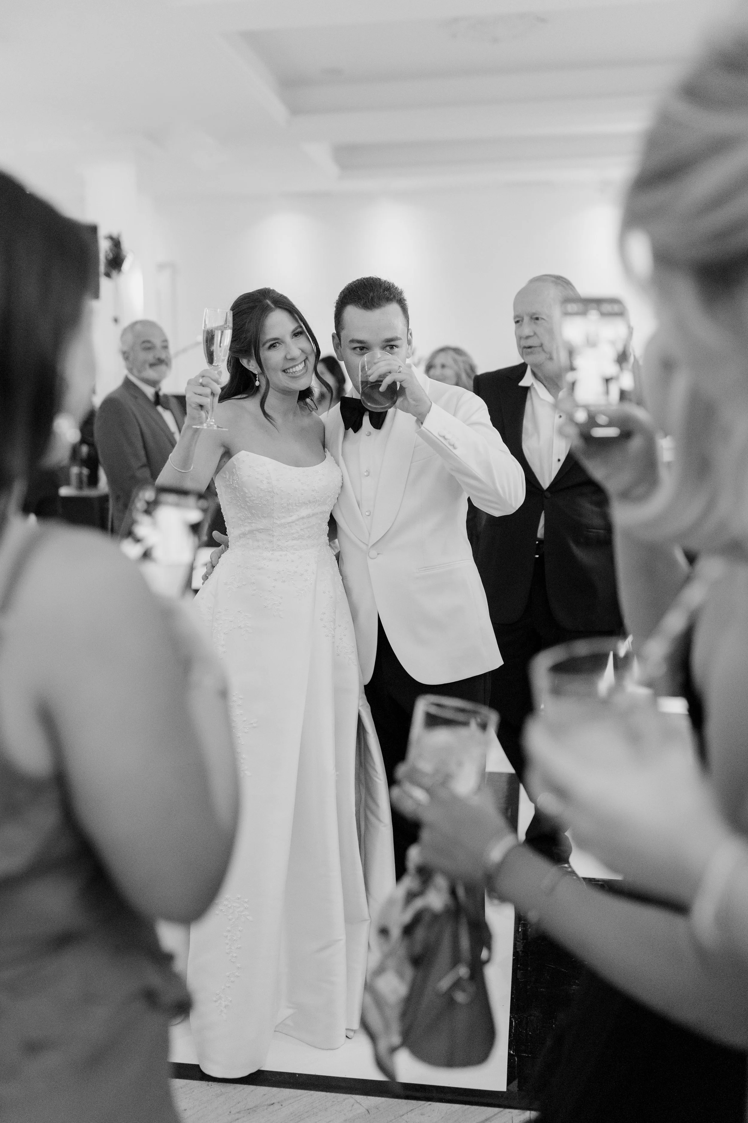 Black and white photo of a wedding celebration with a bride and groom posing for pictures, people in the foreground holding drinks, some smiling, and guests in the background