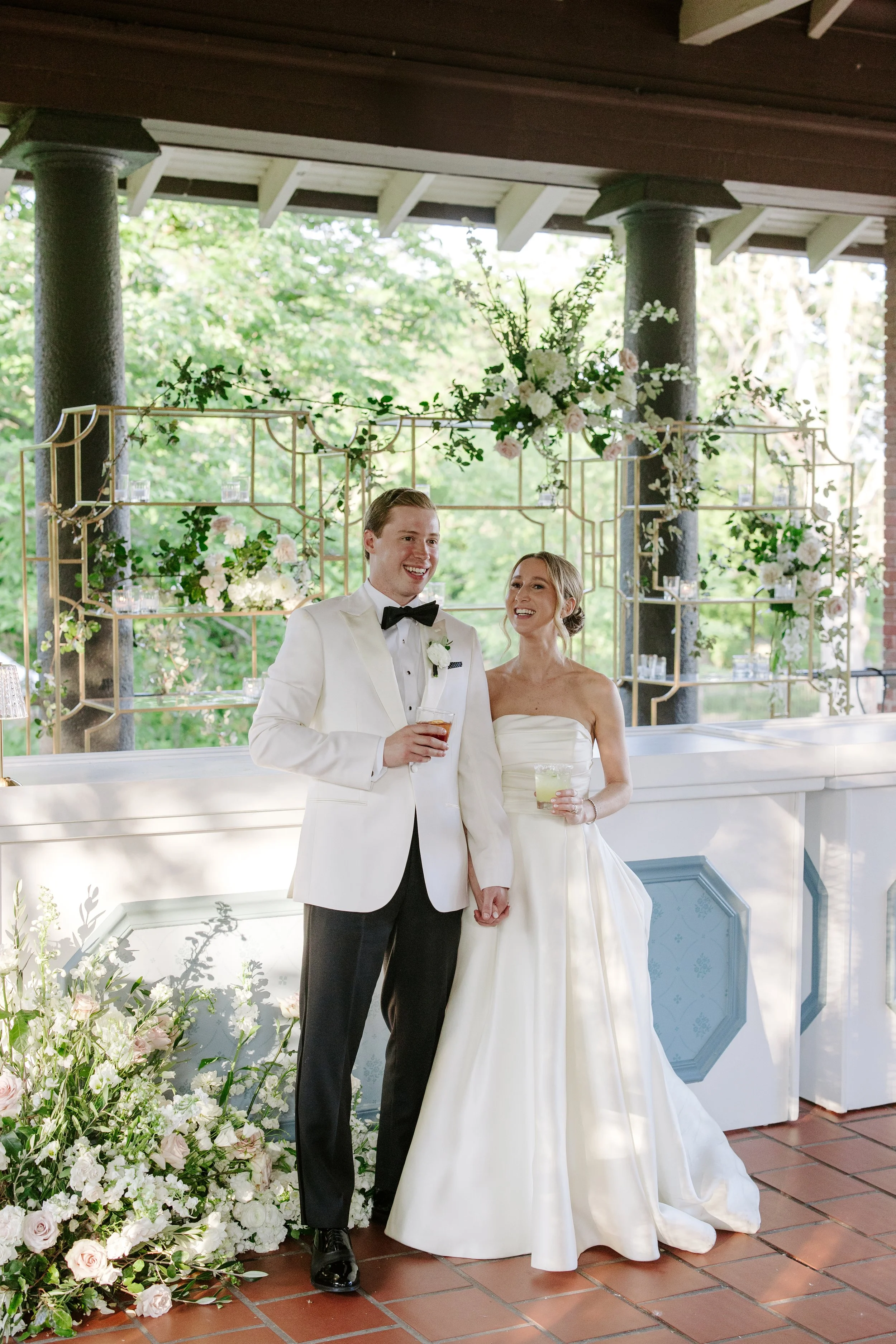 A bride and groom standing together at a wedding reception, holding hands and smiling. The groom is wearing a white tuxedo with a black bow tie, and the bride is in a white strapless wedding dress. They are surrounded by floral arrangements, with a d