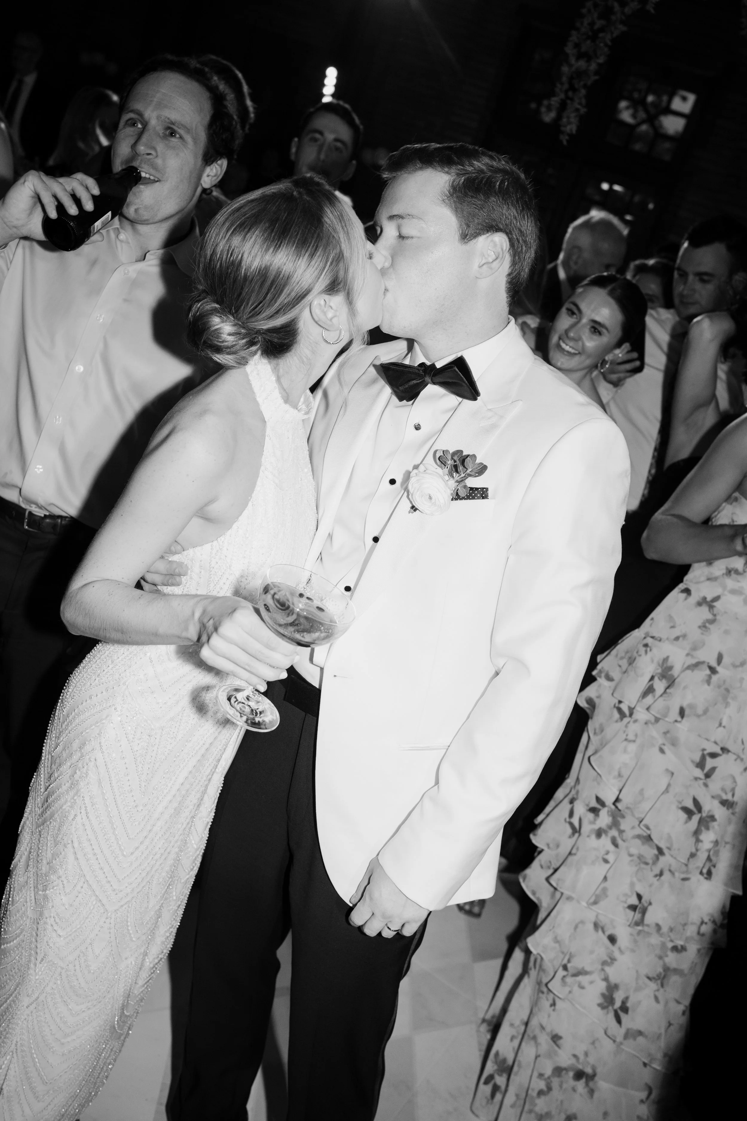 A wedding reception with a bride and groom sharing a kiss, surrounded by smiling guests. The bride is holding a glass of wine, and the groom is dressed in a tuxedo with a bow tie.