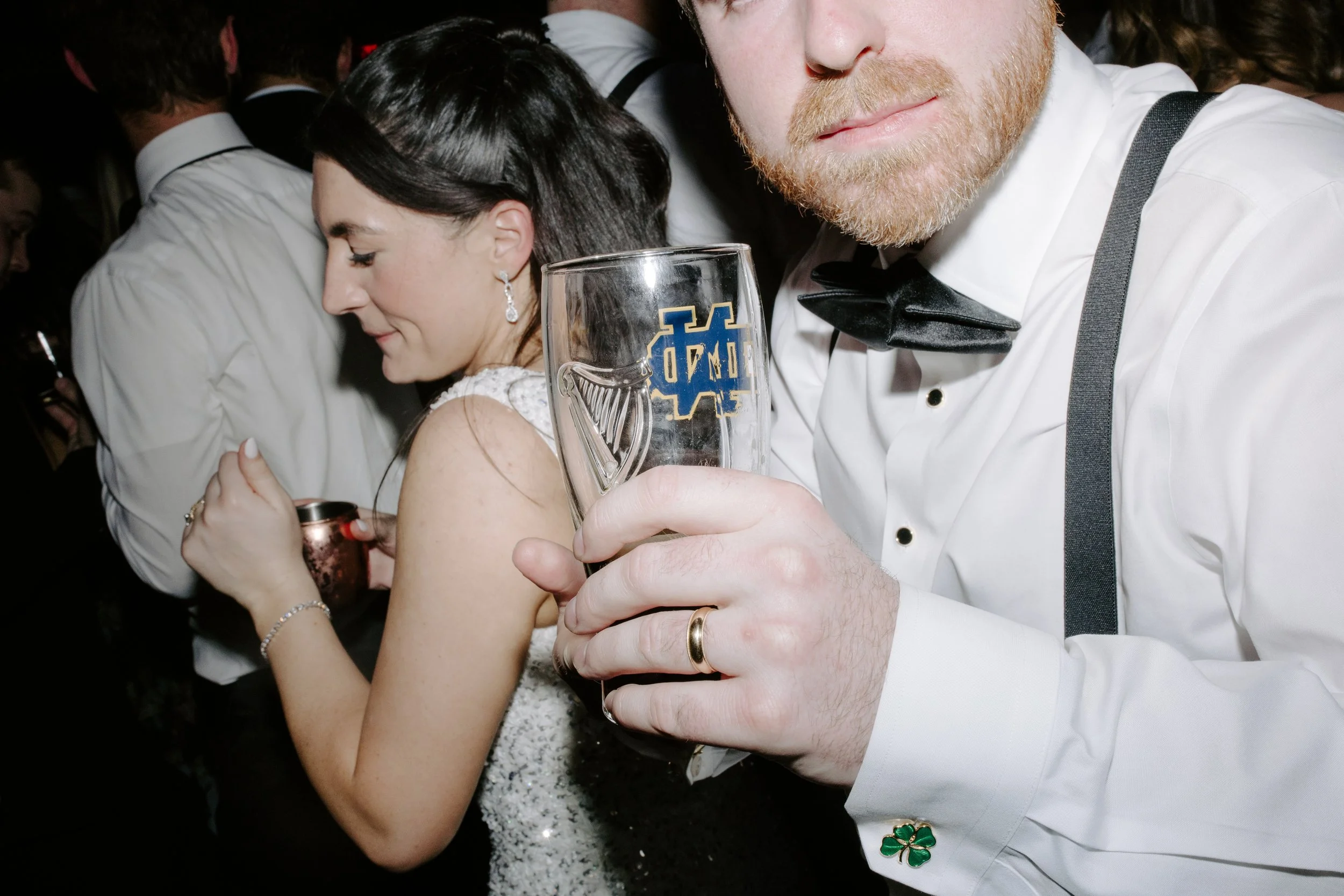 Close-up of a couple at a social event, with the woman wearing a white dress and earrings, and the man in a white shirt with suspenders and a black bow tie, holding a glass of beer with a logo, surrounded by other people.